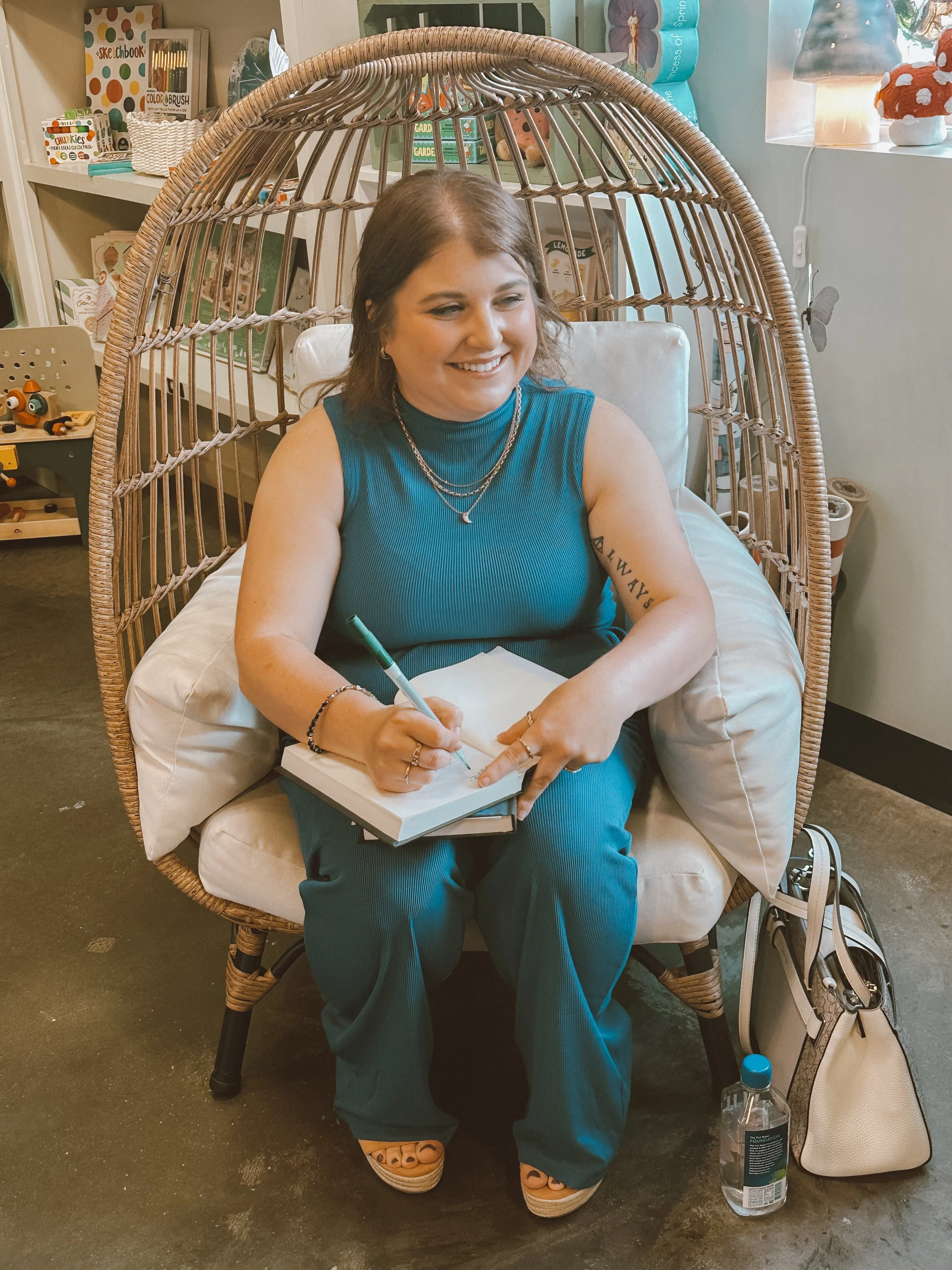 A woman with brown hair smiling, sitting in a wicker hanging chair with cream cushions, writing in a notebook with a green pen, wearing a teal sleeveless top and teal pants, with a beige handbag, water bottle, and shop around her in the background.