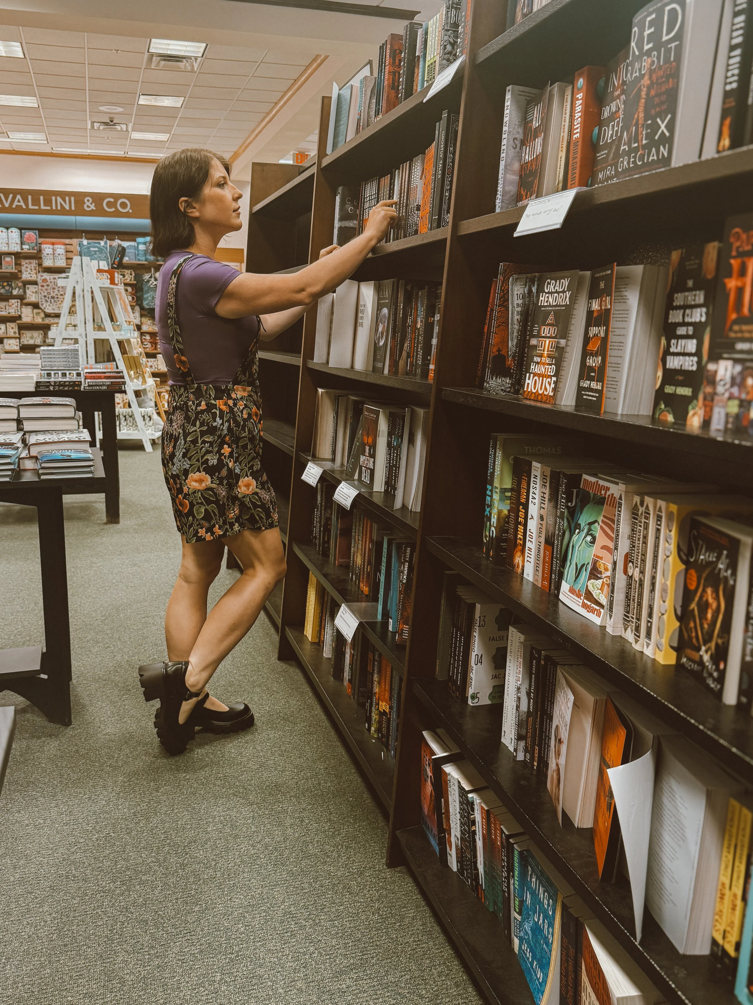A woman in a purple shirt and floral shorts browsing books on a tall bookshelf at a bookstore.