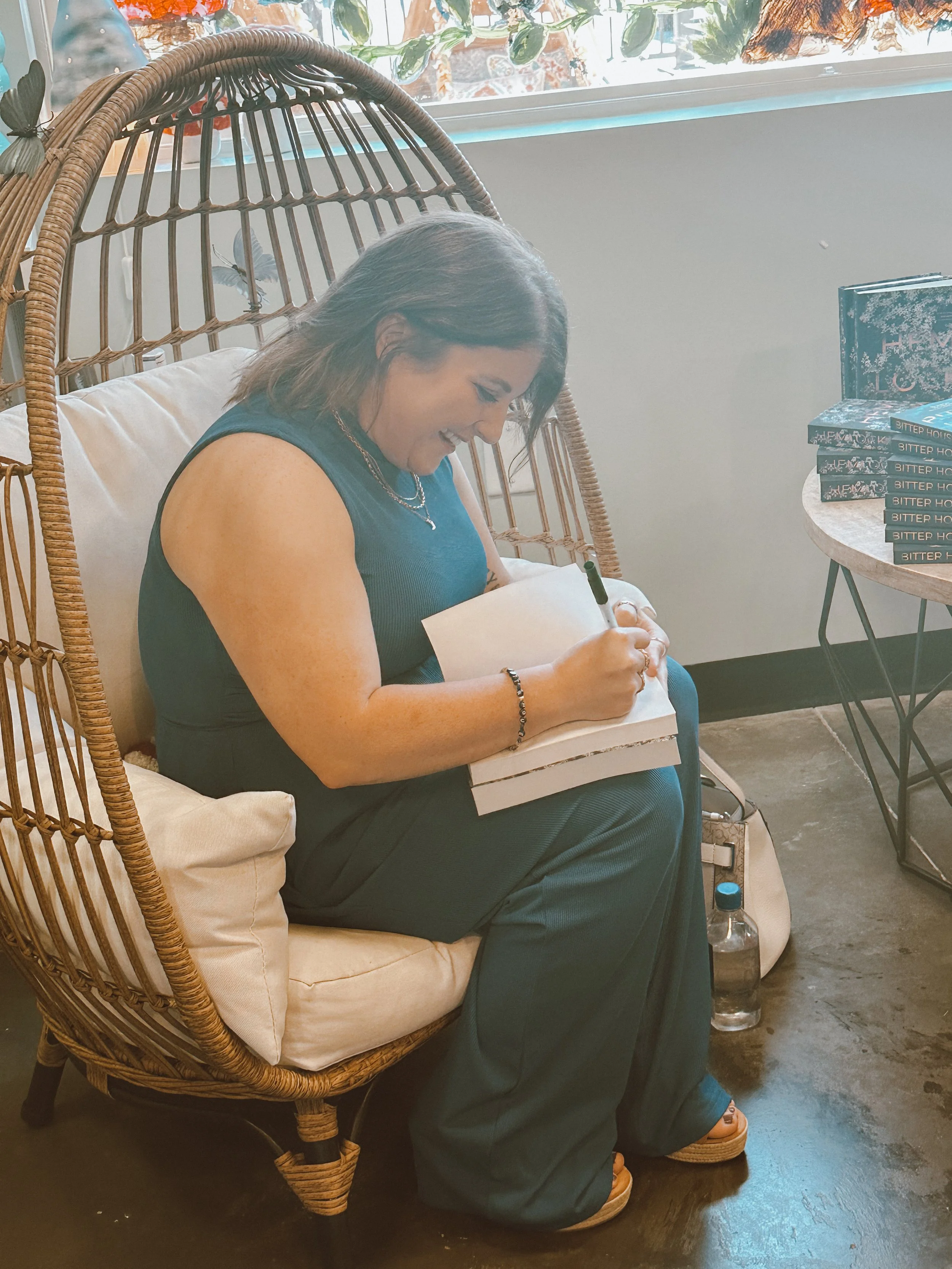 A woman sitting on a wicker chair, smiling and signing a book with a black marker. She is wearing a sleeveless blue dress, jewelry, and sandals. Behind her is a window with plants outside and a small table with stacked books.