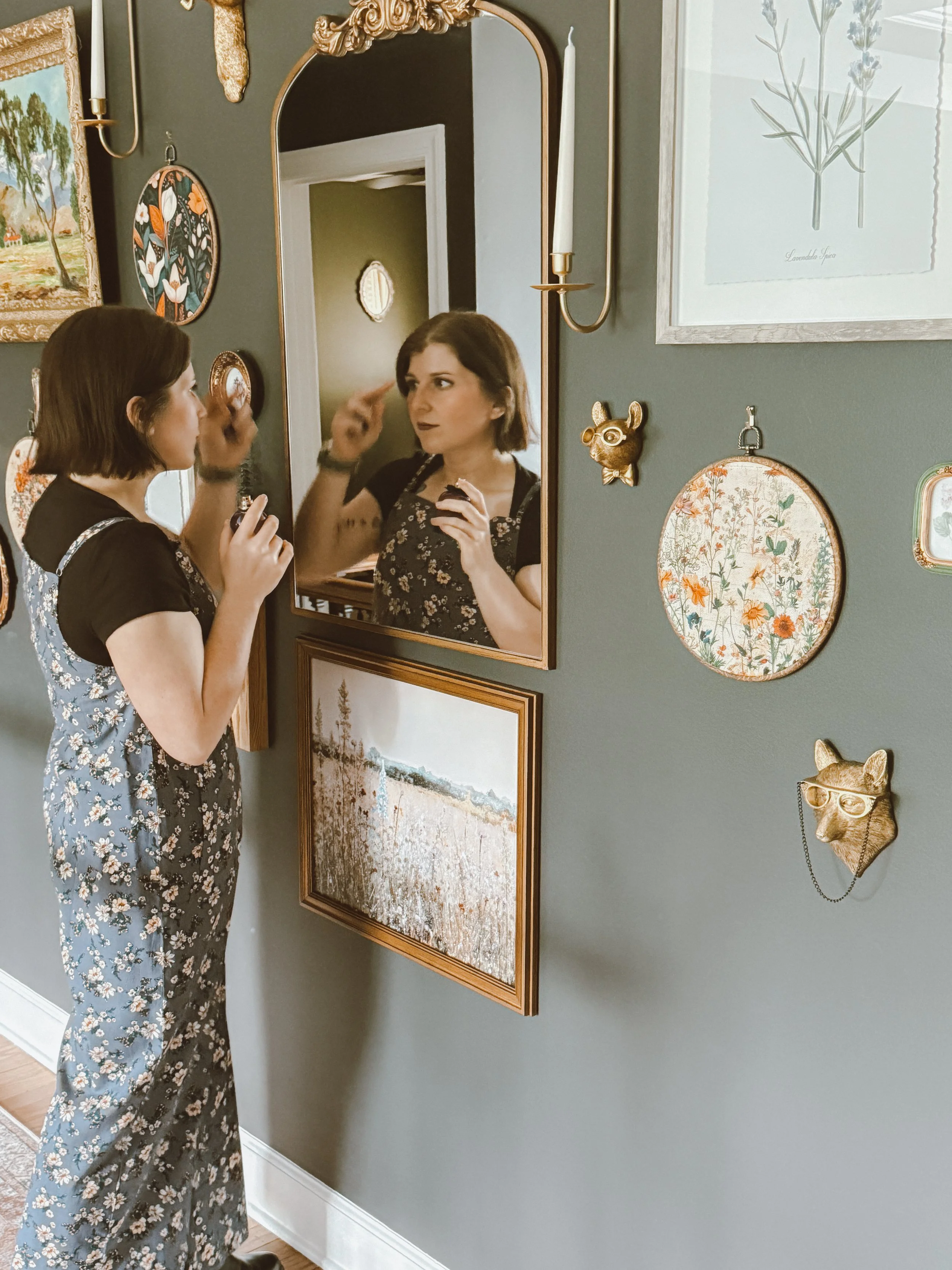 A woman with shoulder-length brown hair wearing floral pajamas is looking at herself in a mirror while applying makeup or skincare. The wall behind her is decorated with framed artwork and small decorative items, including a gold animal face with glasses.
