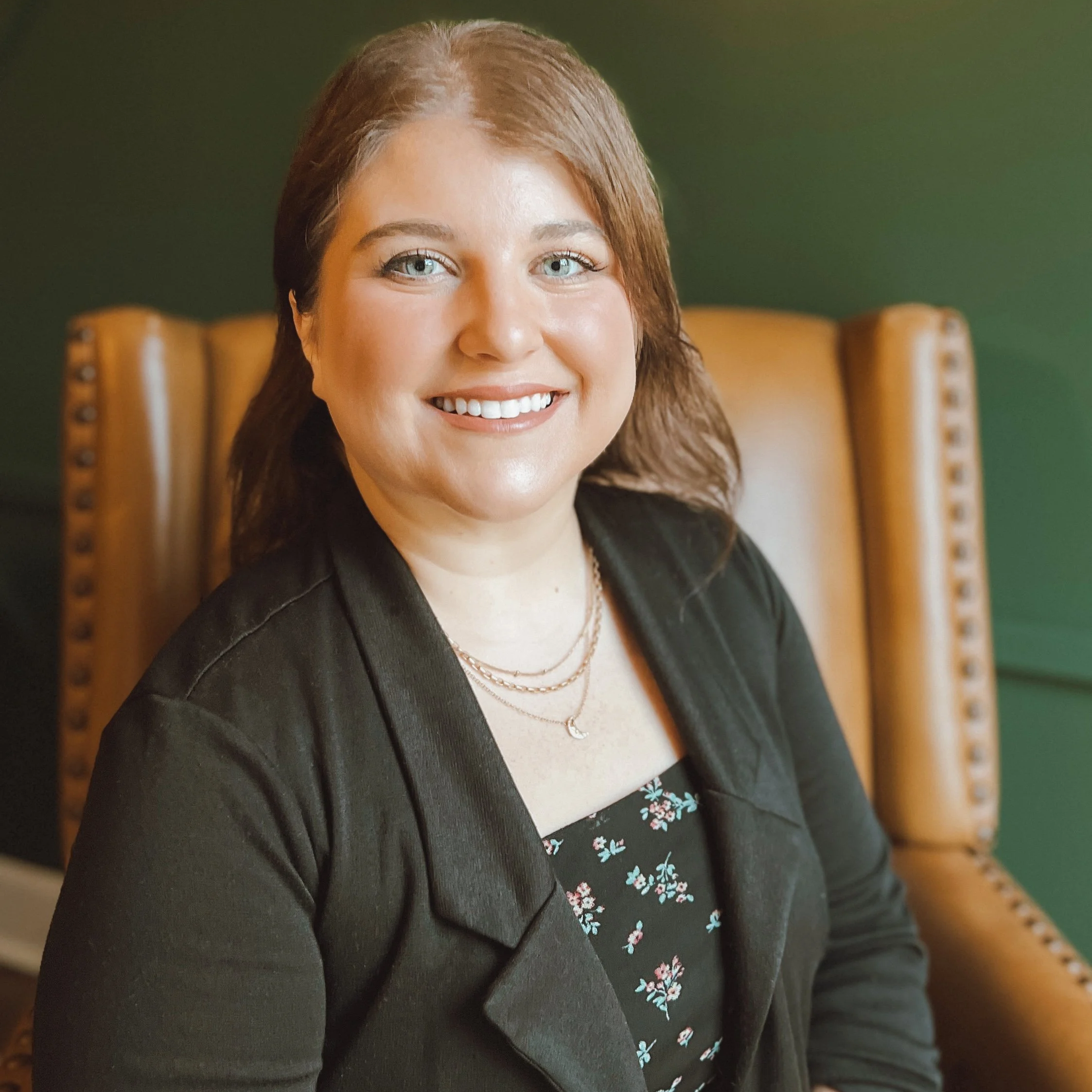 A woman with shoulder-length brown hair and blue eyes smiling, sitting in a tan leather armchair with green background, wearing a black blazer, floral top, and layered necklaces.