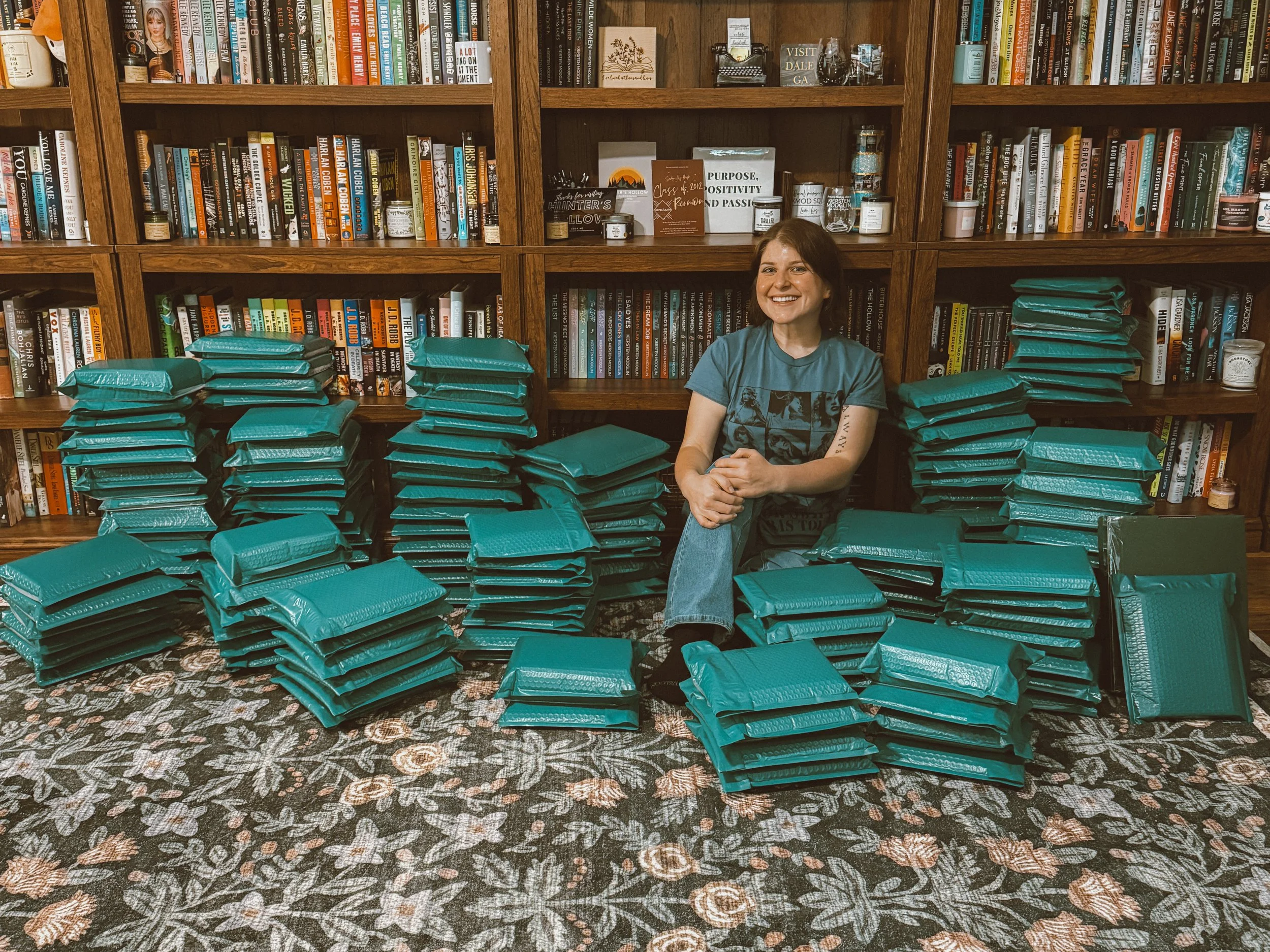A woman smiling and sitting on the floor surrounded by stacks of teal envelopes, in front of a wooden bookshelf filled with books and decor items.