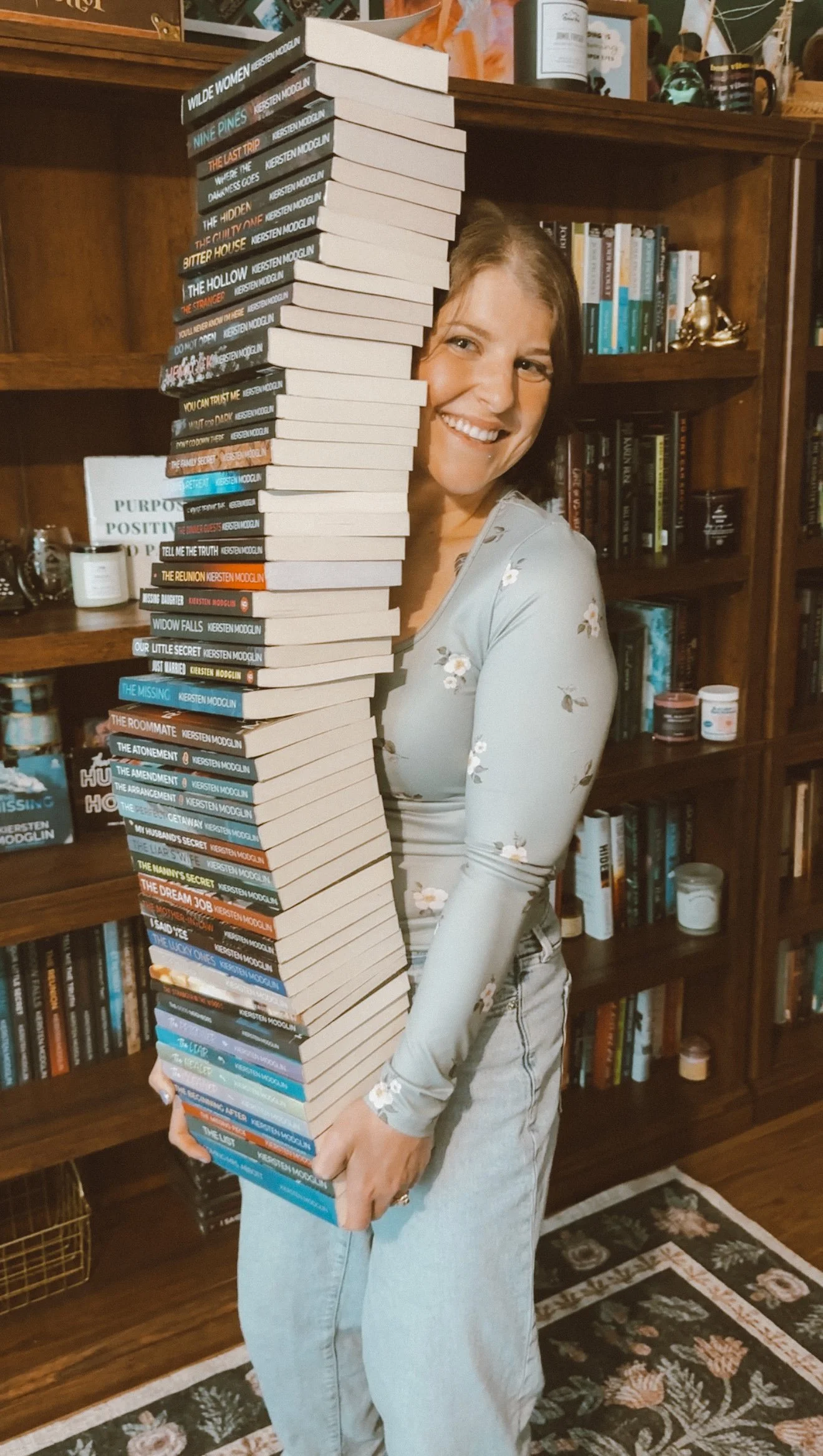 A woman holding a large stack of books indoors with shelves of books in the background.
