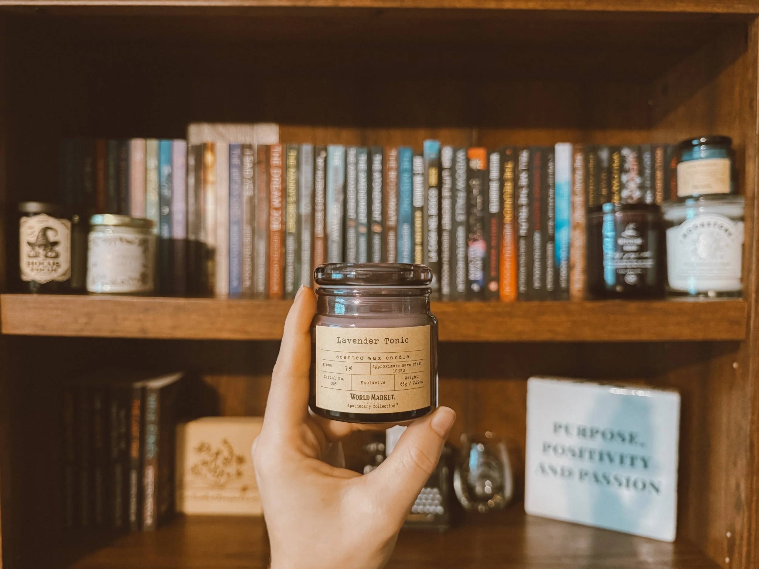 A person's hand holding a lavender scented wax candle with a label reading 'Lavender Tonic' in front of a wooden bookshelf filled with books and jars.