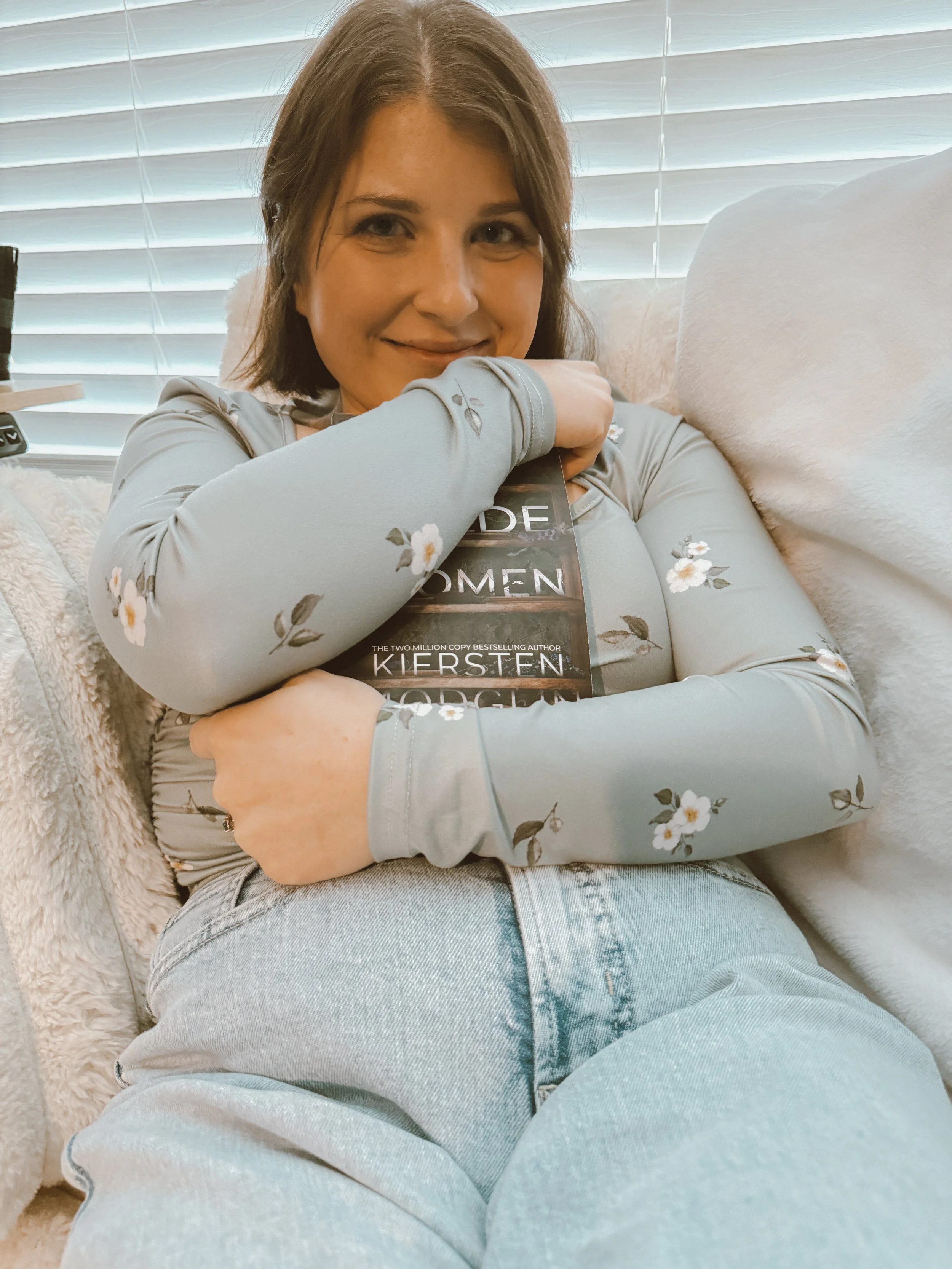 A woman with shoulder-length brown hair, sitting on a beige couch, hugging a book titled 'The Silent Patient' by Alex Michaelides, with a window with blinds in the background.