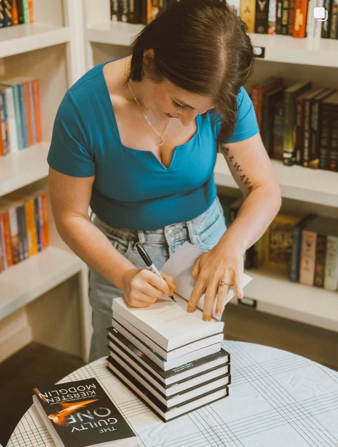 Woman signing a book in a bookstore or library, with shelves of books in the background.