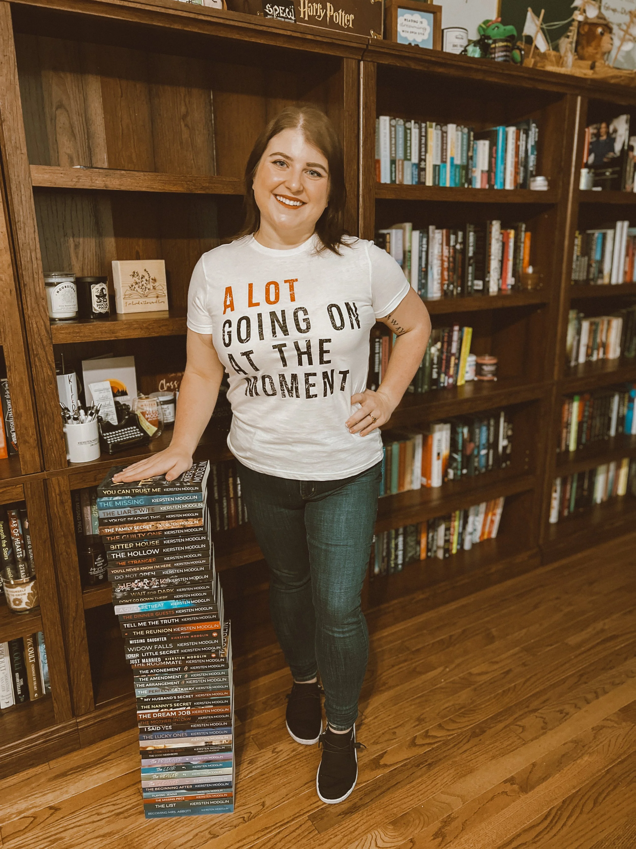 A woman with shoulder-length brown hair, smiling, standing next to a tall stack of books on the floor, in a room with wooden shelves full of books and collectibles.