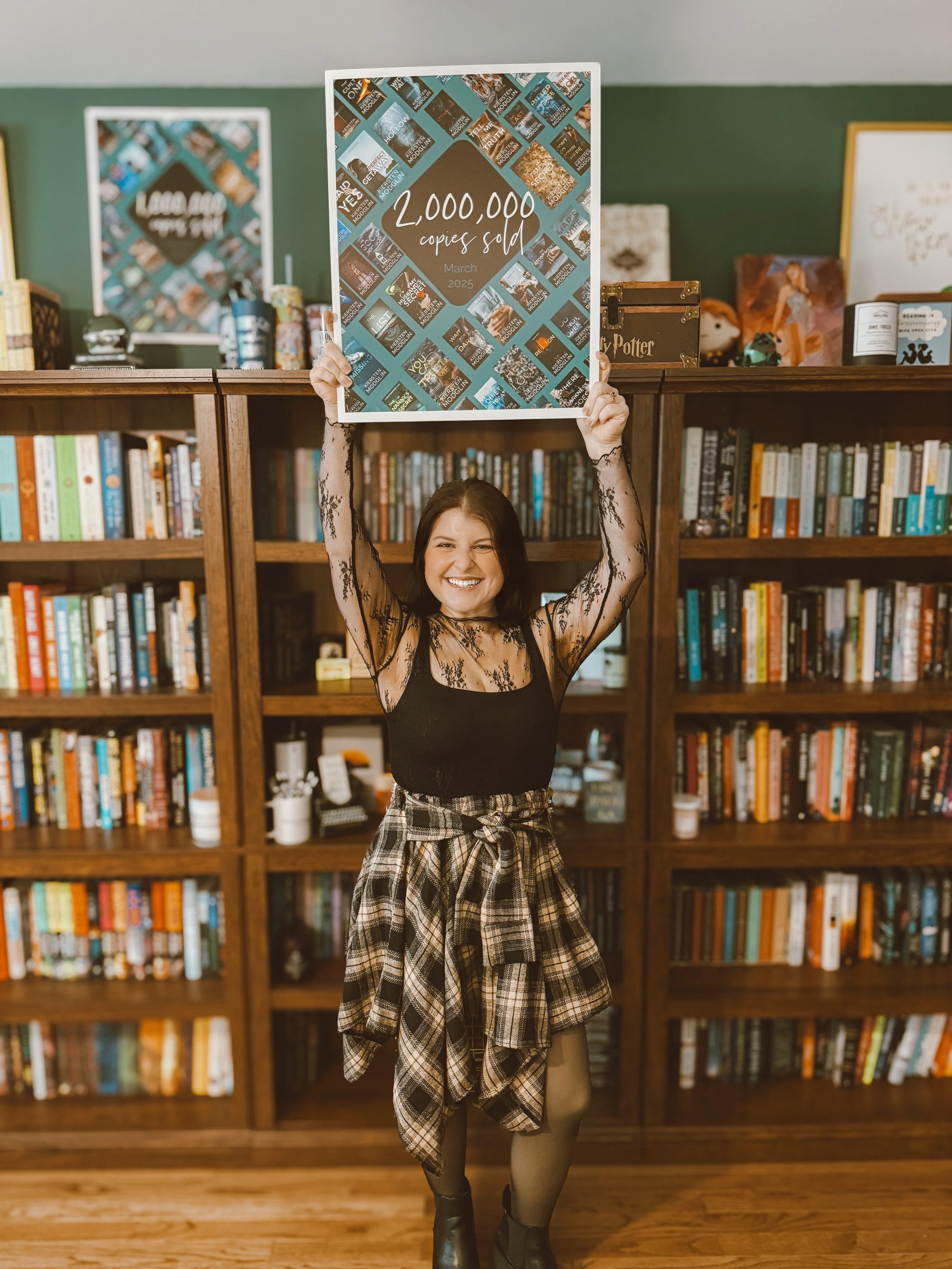 A woman smiling and holding a large sign that reads '2,000,000 copies sold, March 2025' in a bookstore or library with shelves of books in the background.