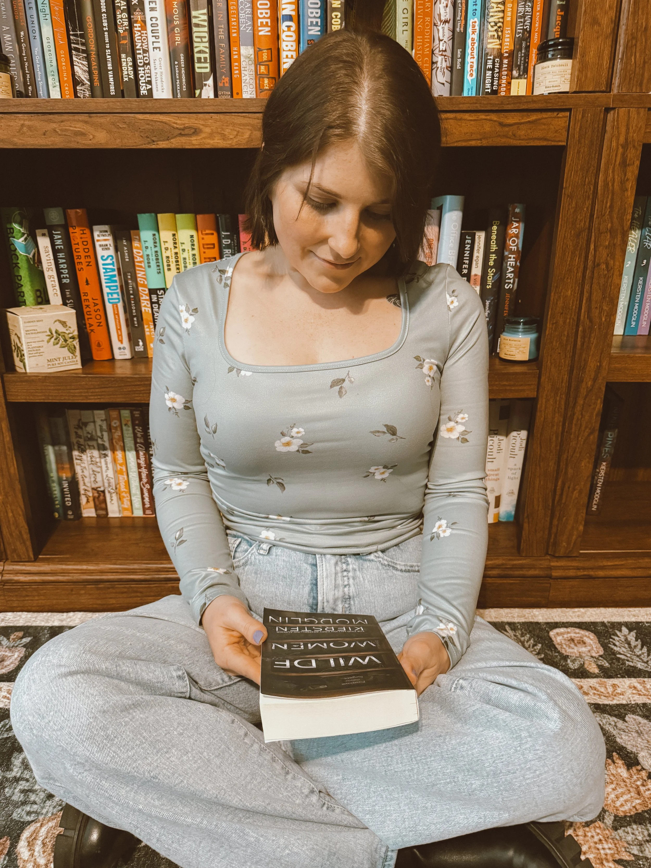 A woman with short brown hair, wearing a light gray floral long-sleeve shirt and light gray jeans, seated cross-legged on a patterned rug in front of a wooden bookshelf, looking down at a paperback book titled 'Wilde Autorennen' in her hands.