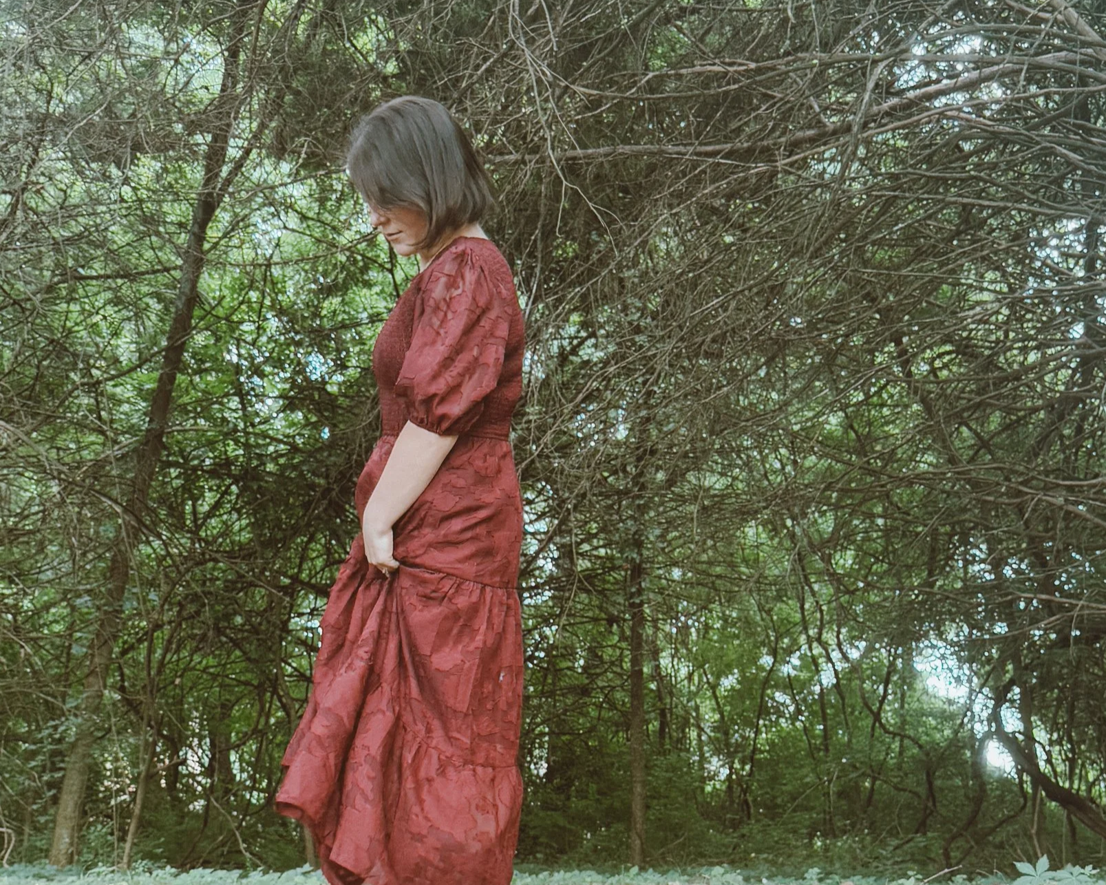 A woman in a long, red, textured dress standing outdoors near trees and bushes, looking down with a gentle expression.