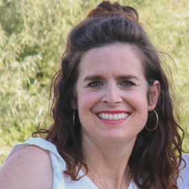 A smiling woman with dark wavy hair in an outdoor setting with trees in the background.