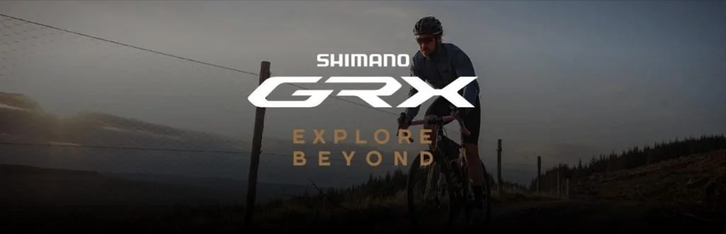 A person riding a mountain bike on a trail near a wire fence, with mountainous terrain in the background, promoting Shimano GRX with the slogan 'Explore Beyond'.