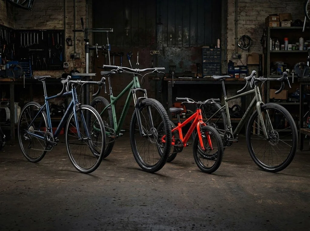 Four bicycles of various sizes and colors, including blue, green, red, and gray, are inside a dimly lit workshop or garage with tools and shelves in the background.