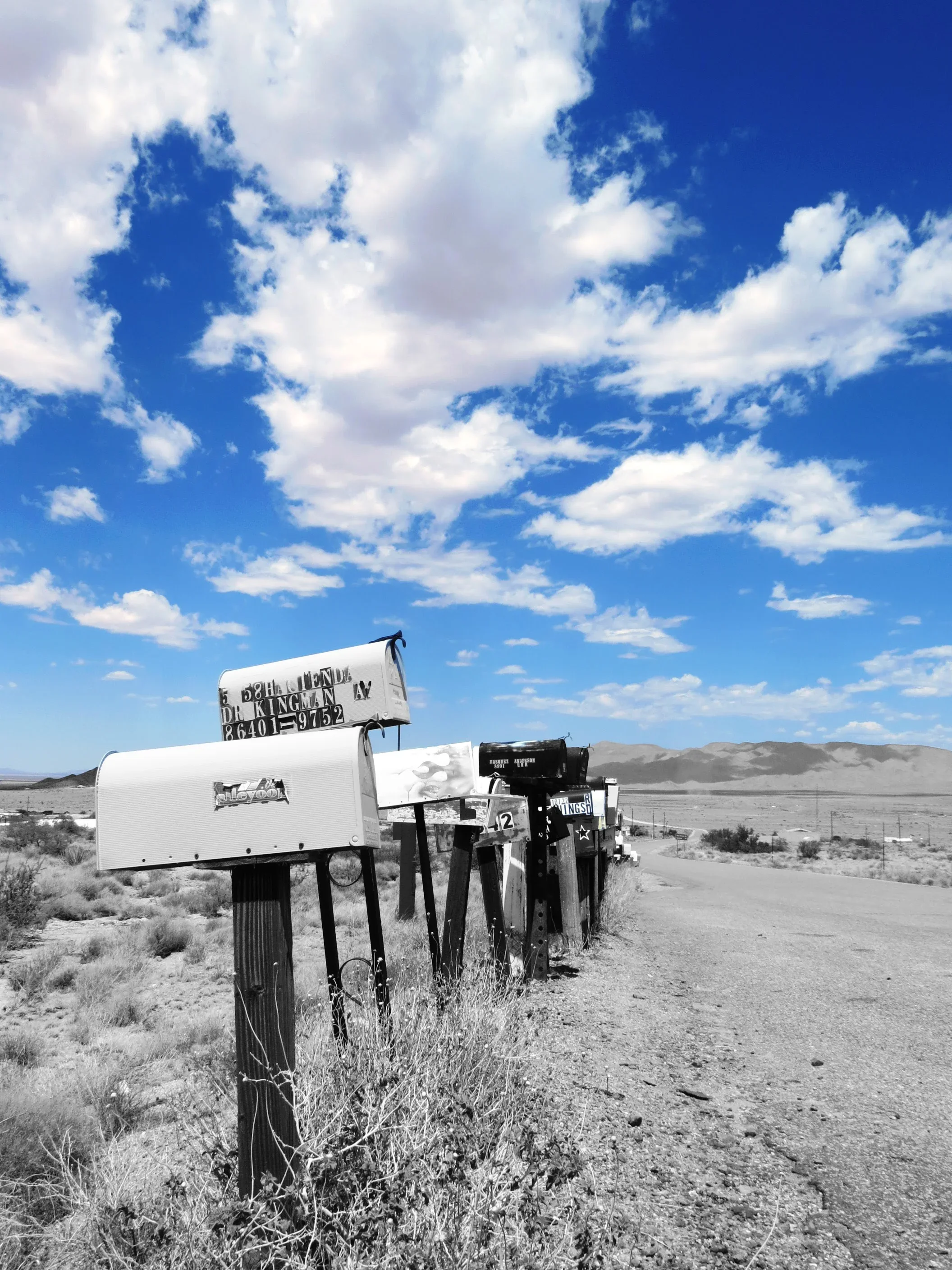A row of mailboxes along a rural dirt road with desert landscape and mountains in the background, under a partly cloudy sky.