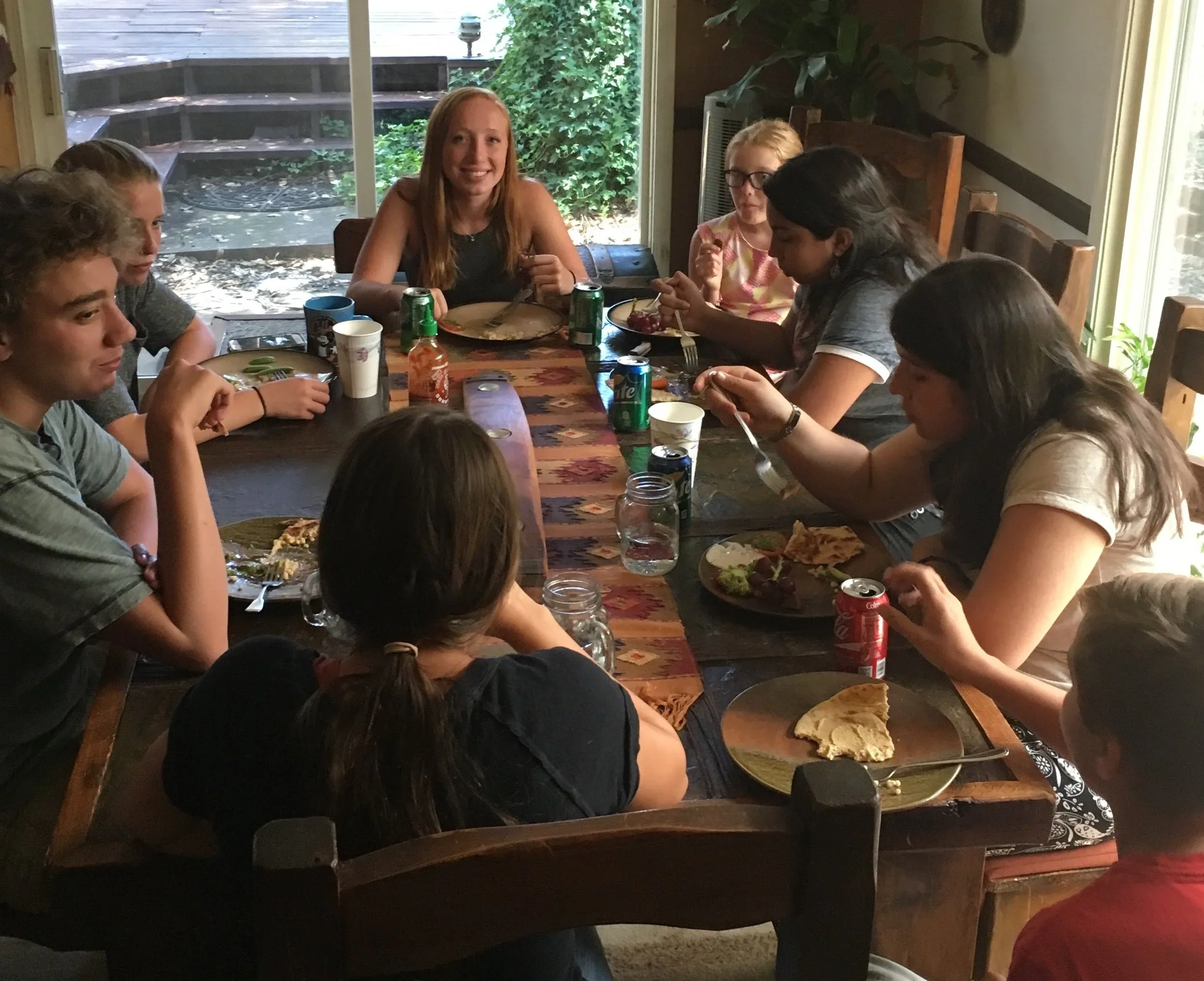 Group of children sitting around a dining table enjoying a meal together, with some food, drinks, and soda cans on the table.