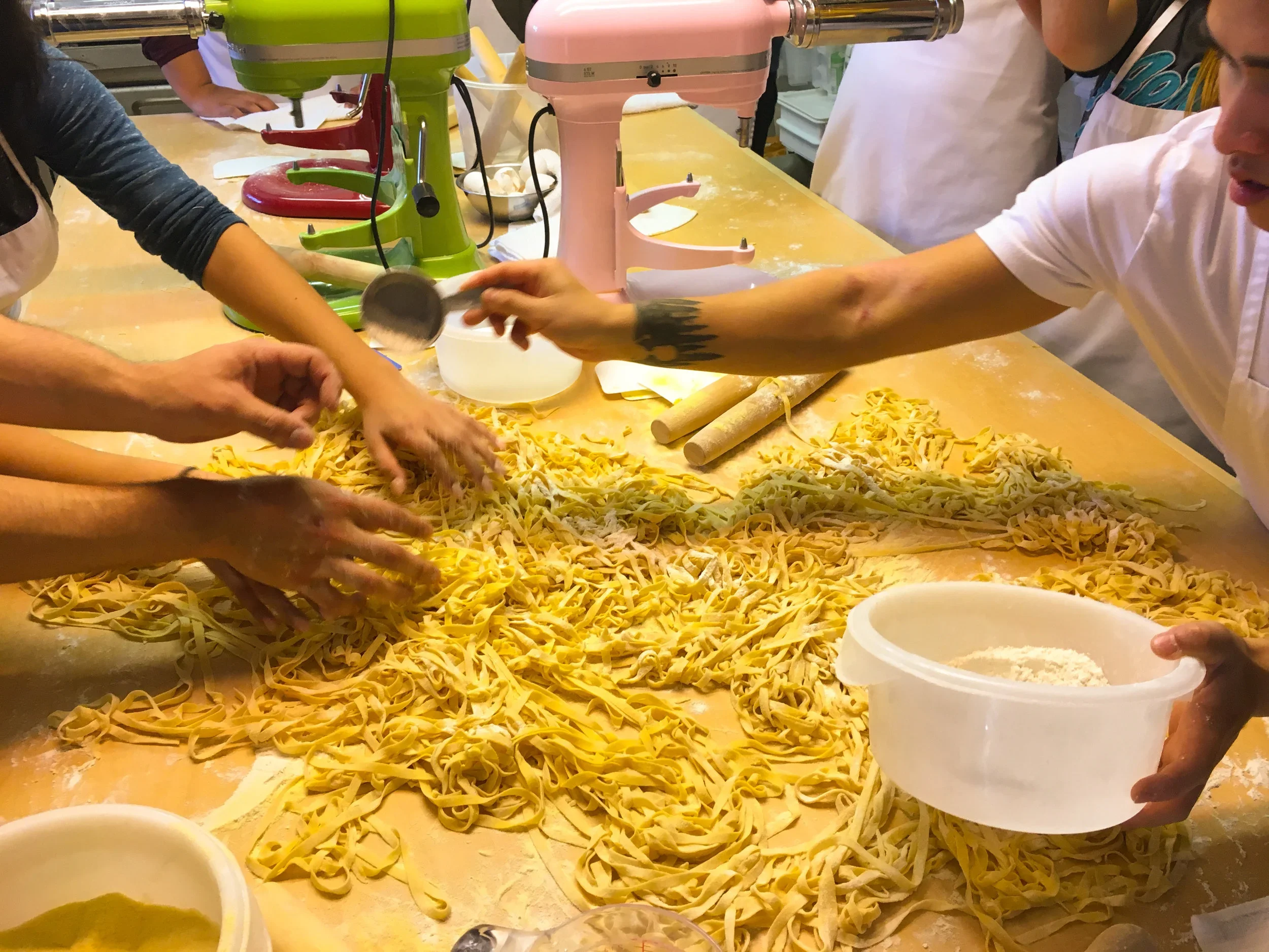 People making pasta dough and cutting into noodles using a pasta machine in a busy kitchen.