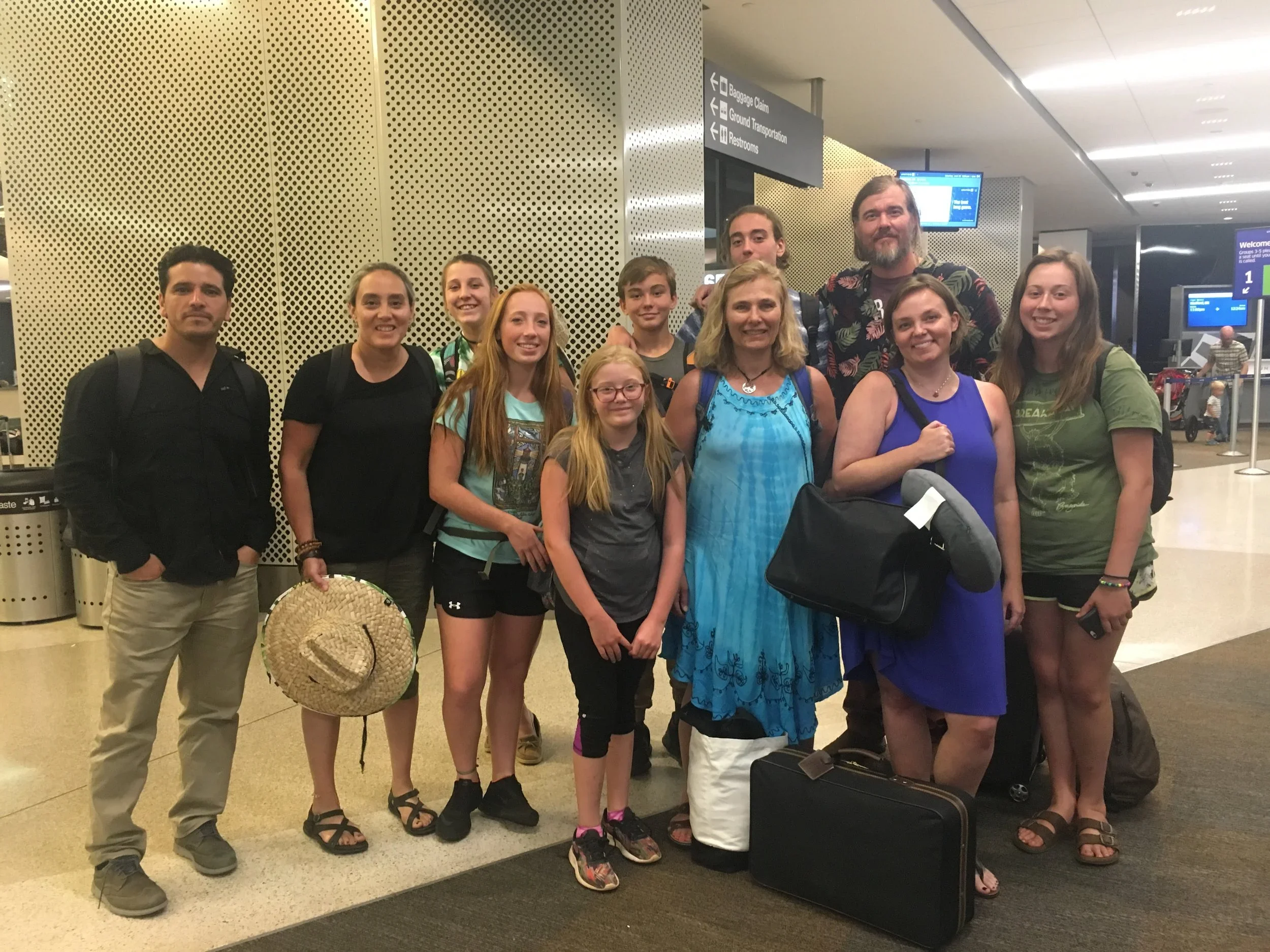 Group of eleven people, including children and adults, standing together at an airport baggage claim area.