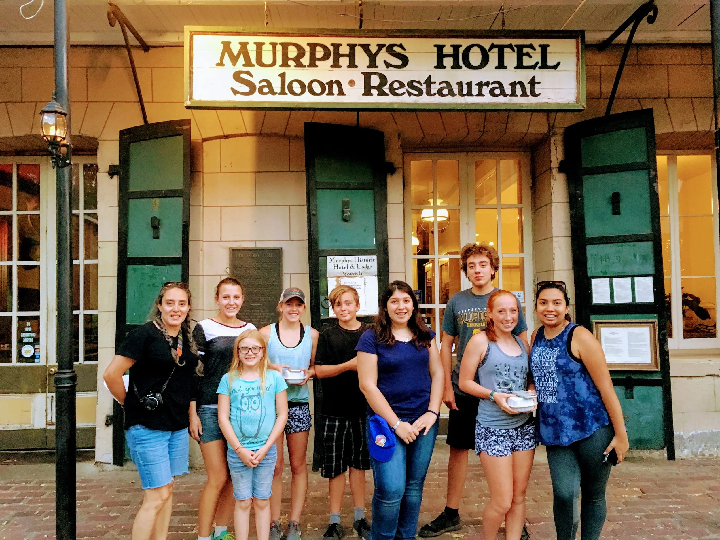 Group of nine people standing outside the Murphys Hotel, which has a sign that reads 'Saloon Restaurant'. The group includes young girls and teenagers, some holding small containers, standing on brick pavement in front of the hotel's entrance.