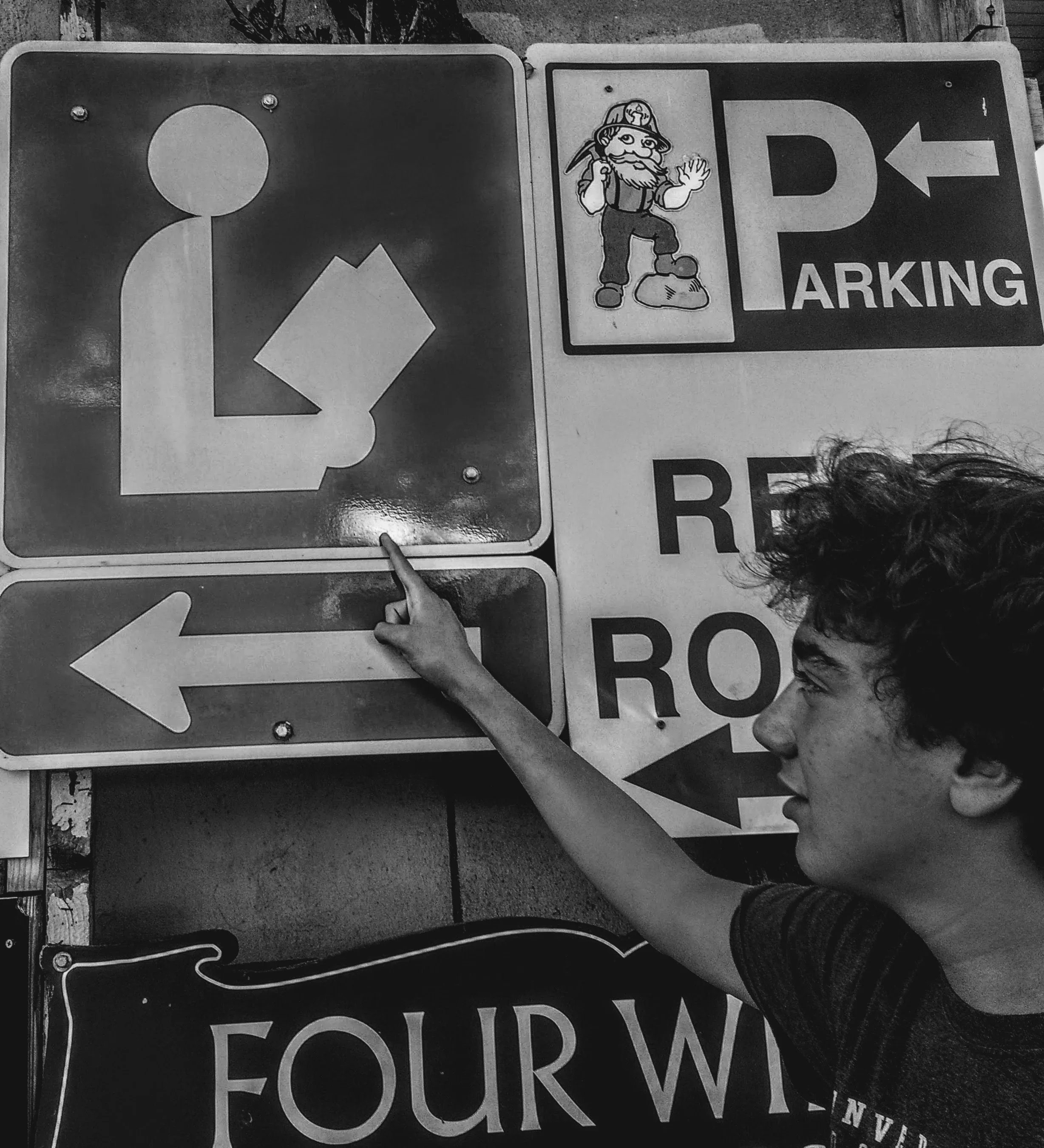 A young person with curly hair pointing to a traffic sign that indicates a left turn. The sign features an arrow pointing left, and the background includes other traffic signs, including a parking sign with a cartoon gnome figure.