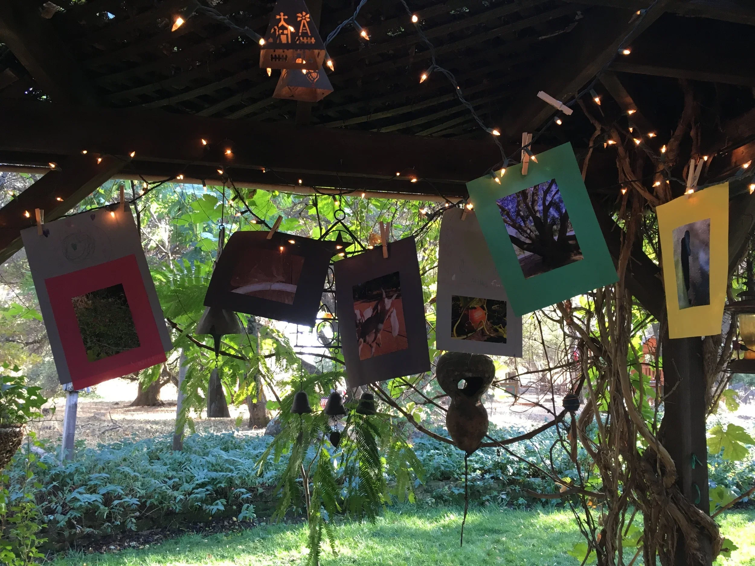 Colorful paper frames with photos hanging from a string in a garden setting, decorated with string lights, under a wooden roof.