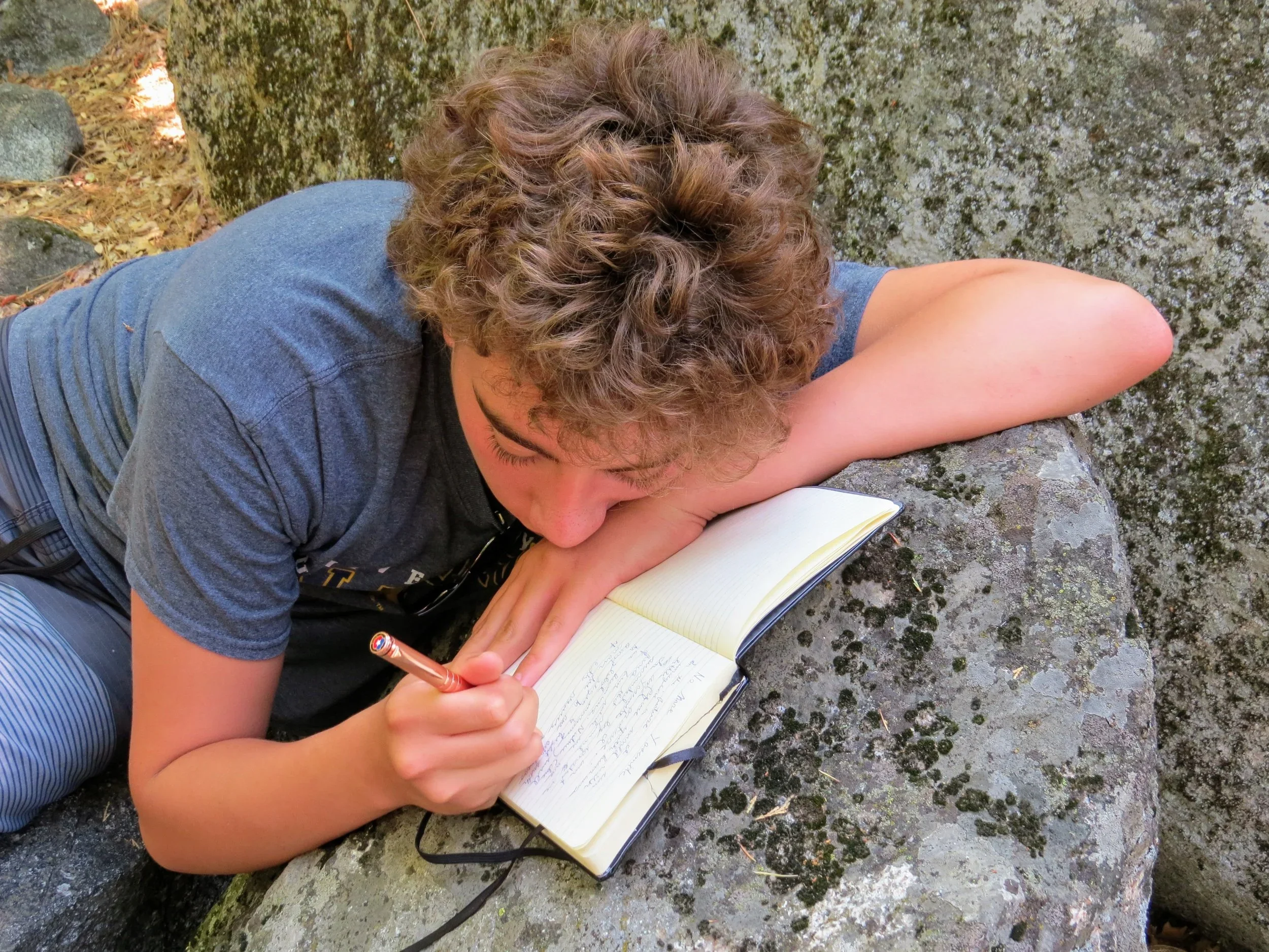 A young person with curly hair lying on a large rock outdoors, writing in a notebook with a pen.