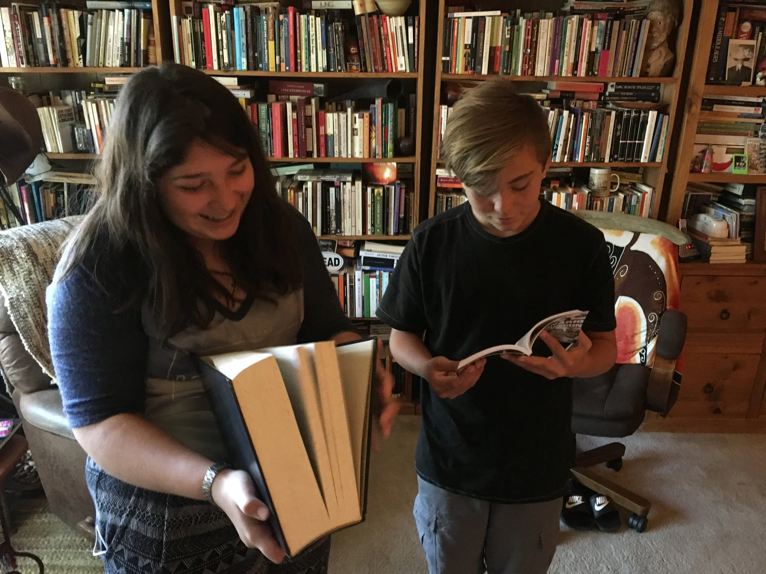 A woman and a boy are reading books in a room with wooden bookshelves filled with books. The woman is smiling and holding a large, open book. The boy is focused on reading a book. There is a chair and some shoes visible in the room.