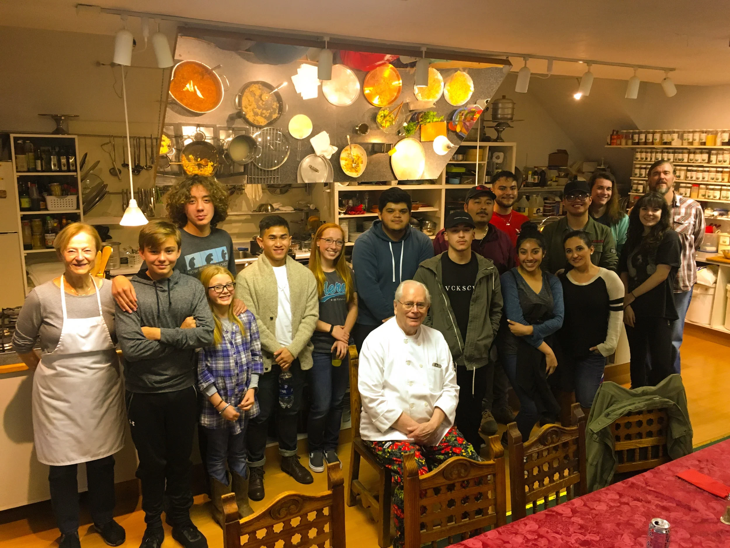 Group of people in a kitchen, including a chef in a white coat and colorful pants, posing for a photo.