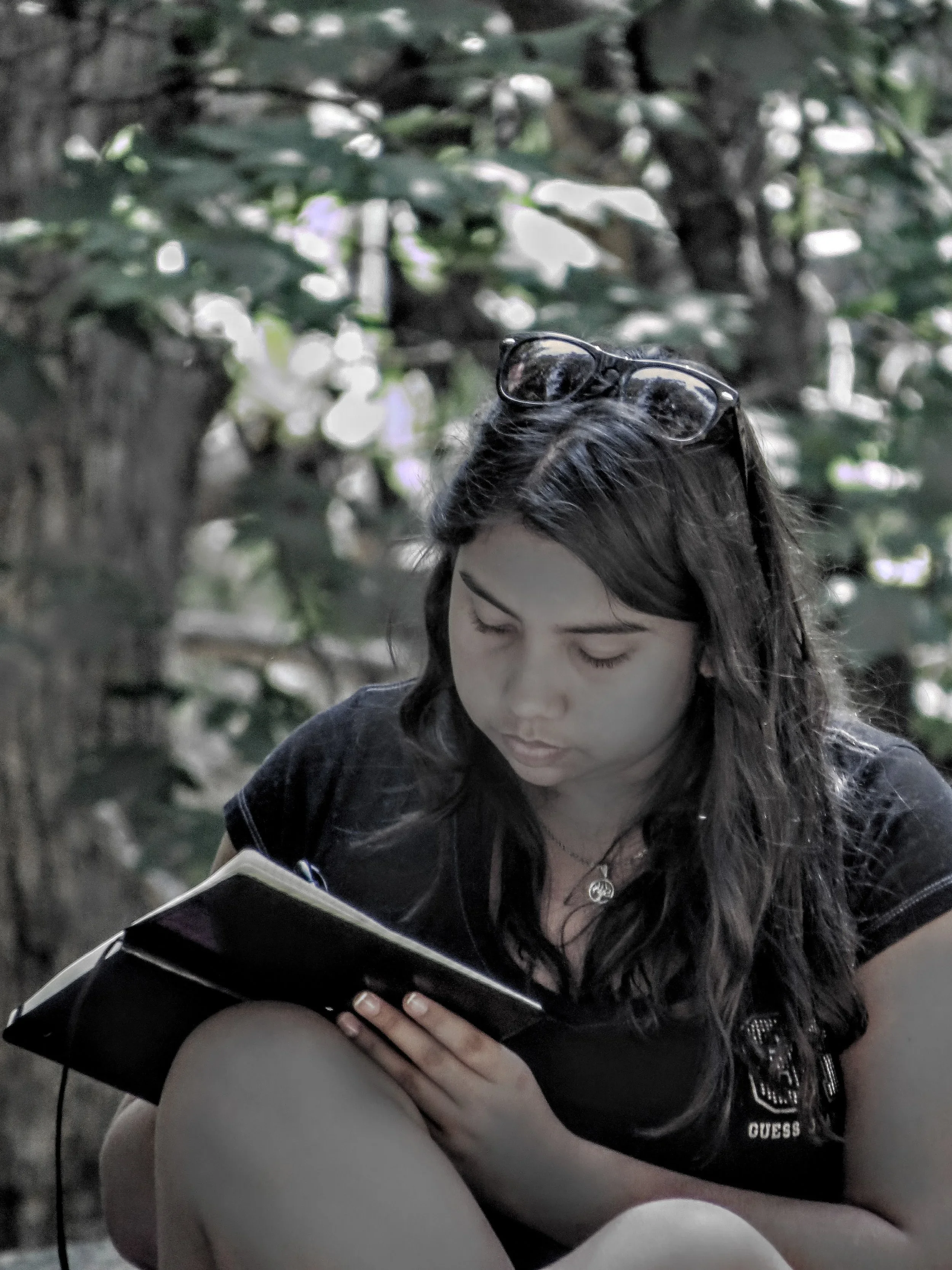 A young woman with long dark hair, glasses on her head, wearing a black t-shirt, is sitting outdoors and reading a black notebook.