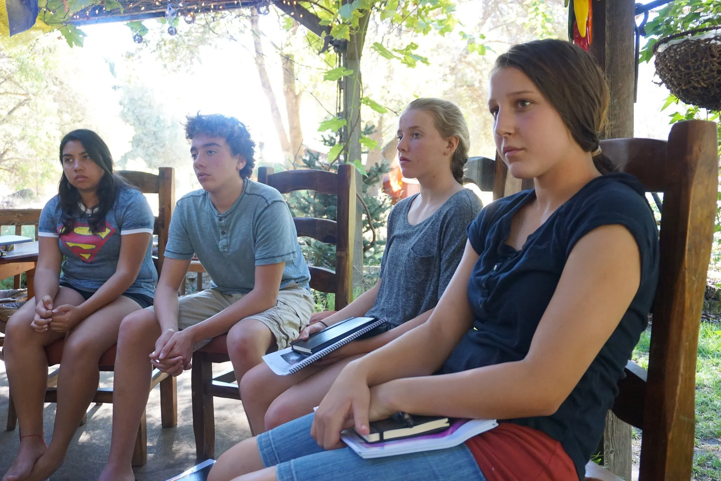 Four young girls sitting on chairs outdoors during daytime, listening attentively, with trees and greenery in the background.