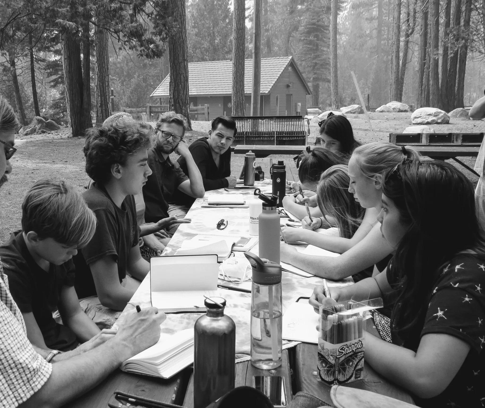 A group of children and teenagers sitting around a long outdoor table in a wooded, outdoor setting, taking notes and listening to a person at the head of the table.
