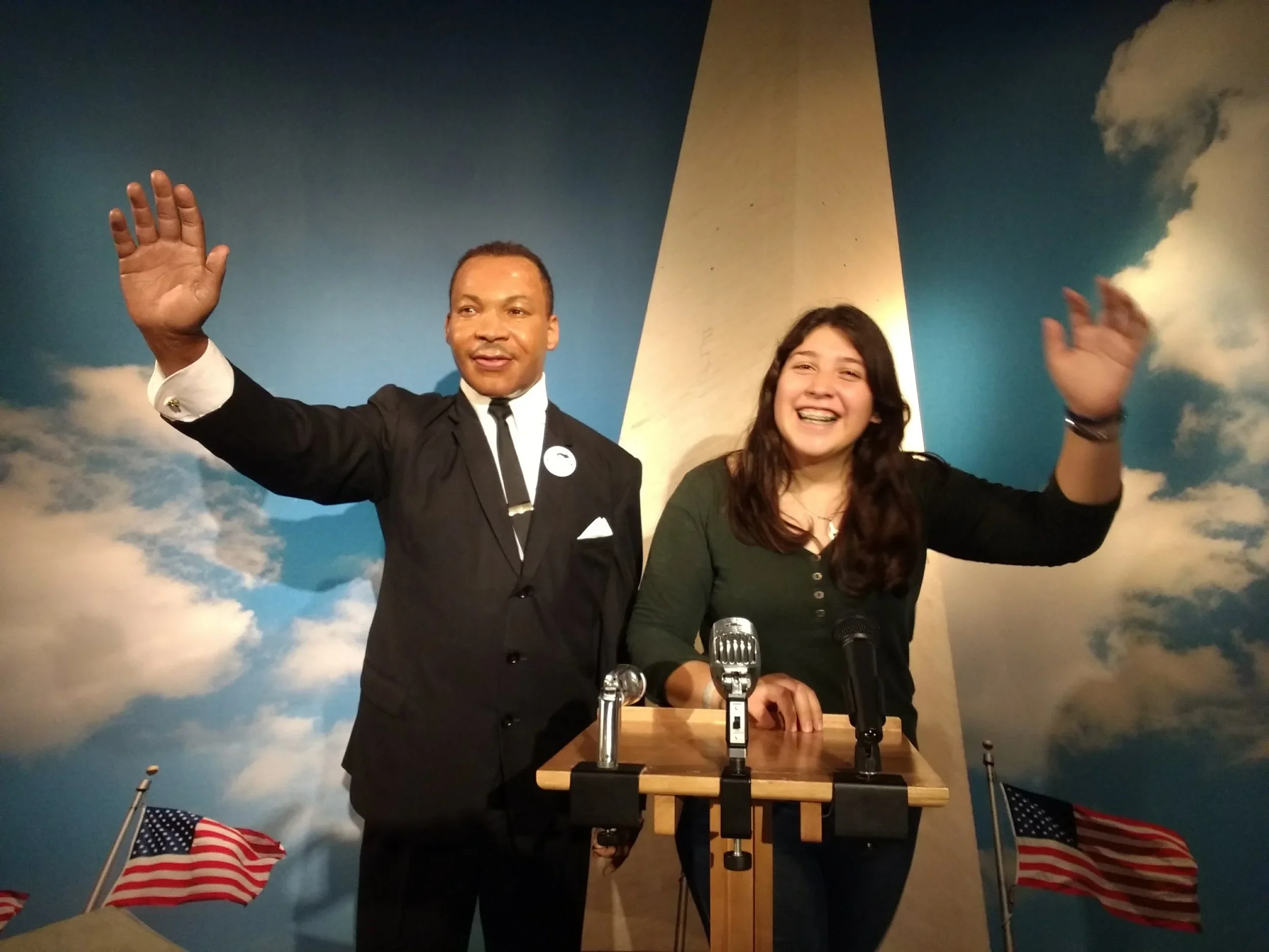 A man in a suit and a young woman are standing behind a podium with microphones, smiling and waving. Behind them is a painted sky with clouds and a tall monument, with American flags at the bottom.