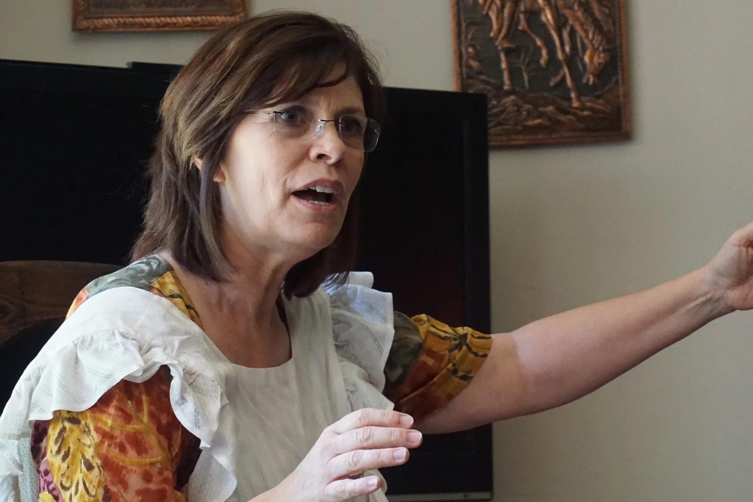 A woman with shoulder-length brown hair, glasses, and a multi-colored dress is raising her right arm and speaking, with artwork hanging on the wall behind her.