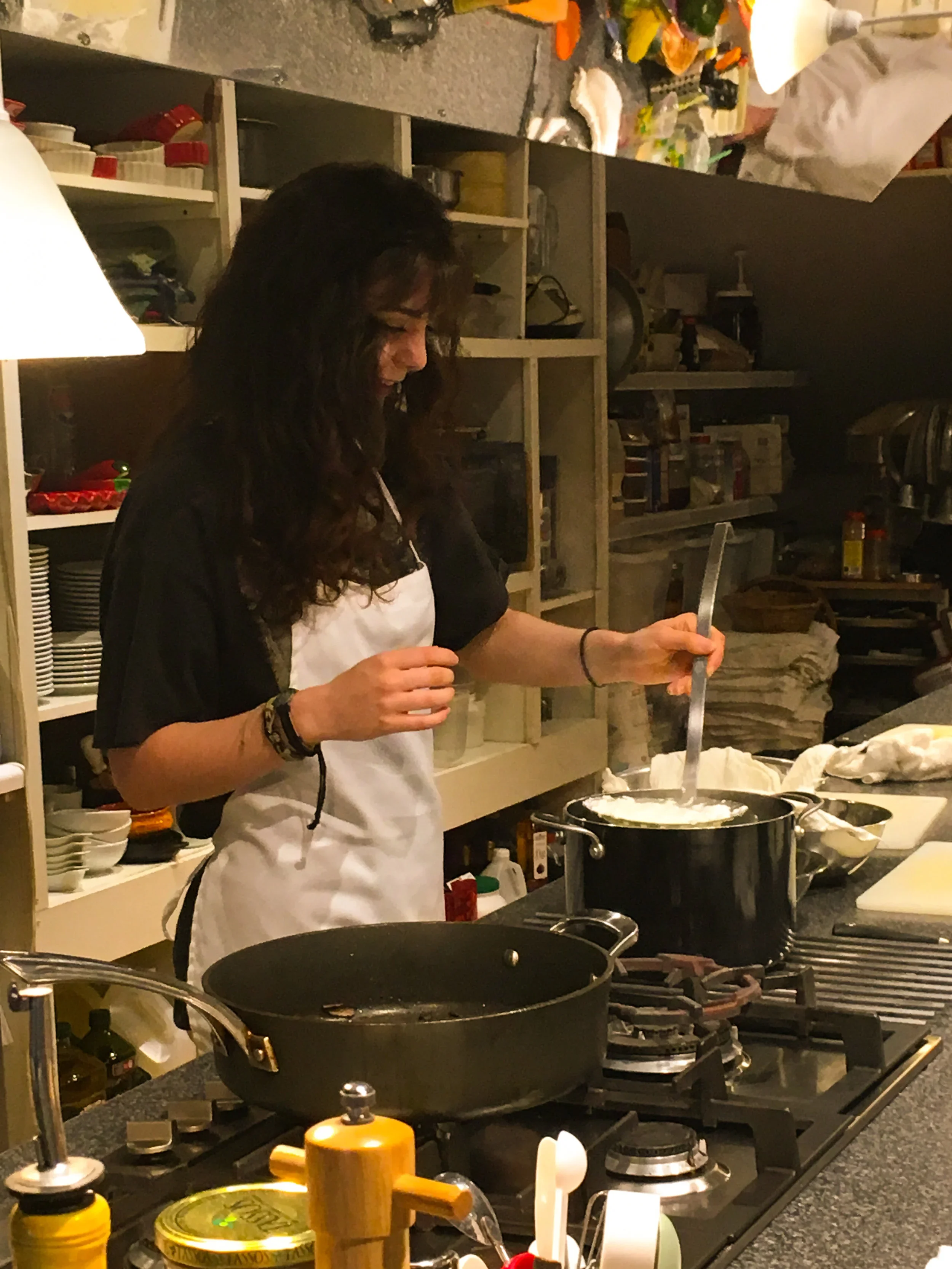 A woman wearing a white apron is cooking on a stove in a kitchen, stirring a pot with a spoon.