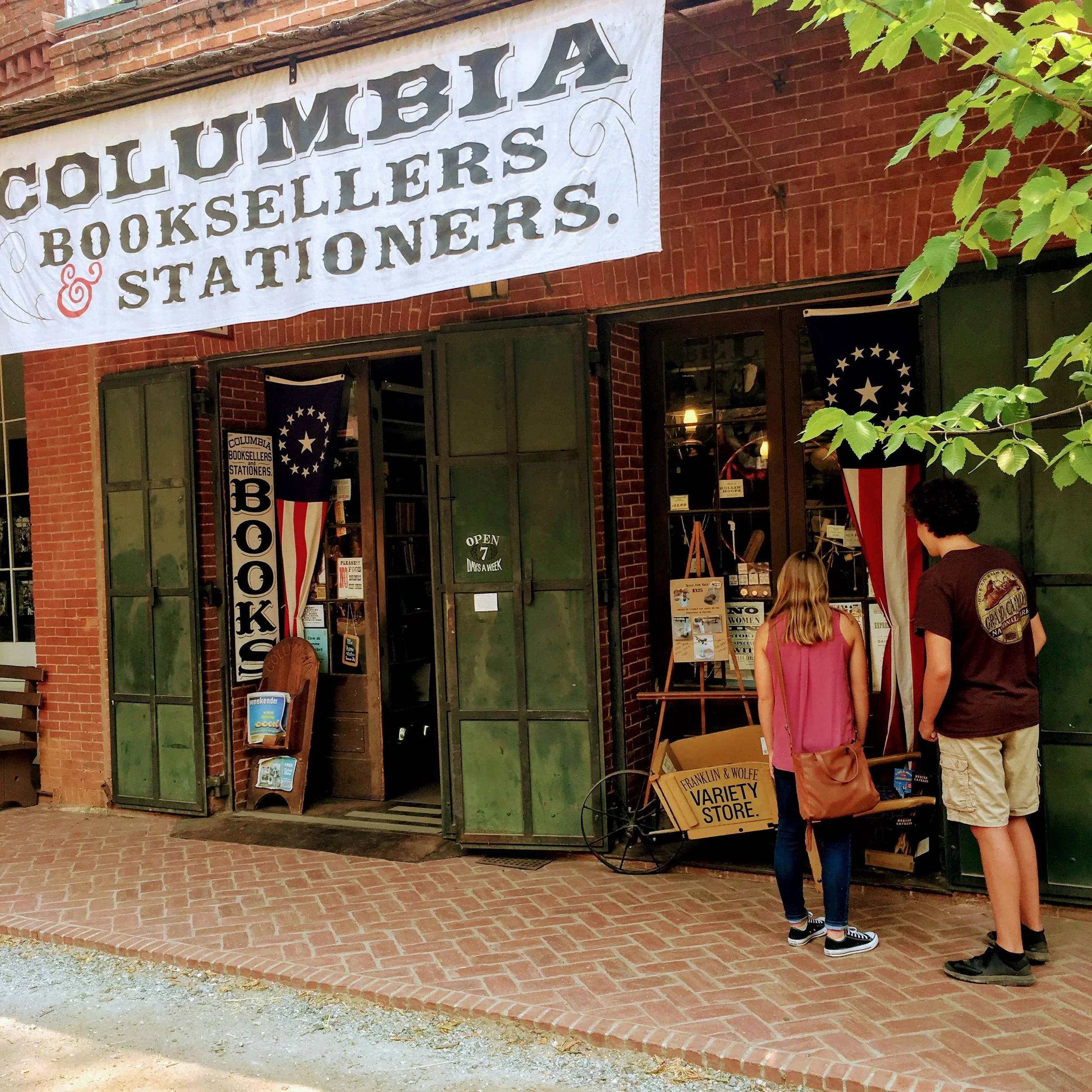 People outside a bookstore with a large sign that reads 'Columbia Booksellers & Stationers.' The storefront has open green shutters and flag banners with stars and stripes. A couple is looking at items displays near the entrance.