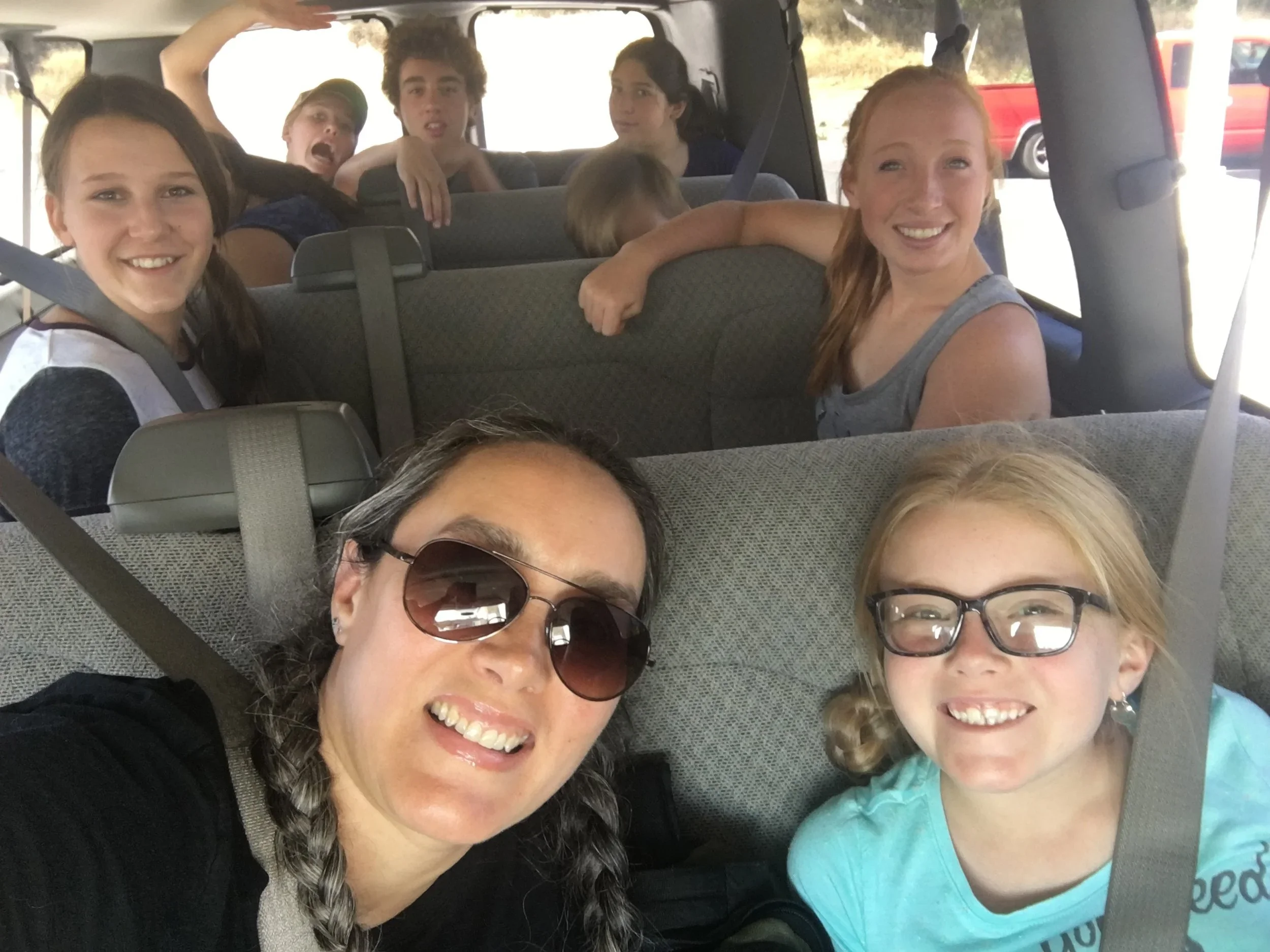 A group of people smiling and sitting inside a van, with several children and two women in the foreground, one wearing sunglasses and another with glasses, taken during a daytime trip.