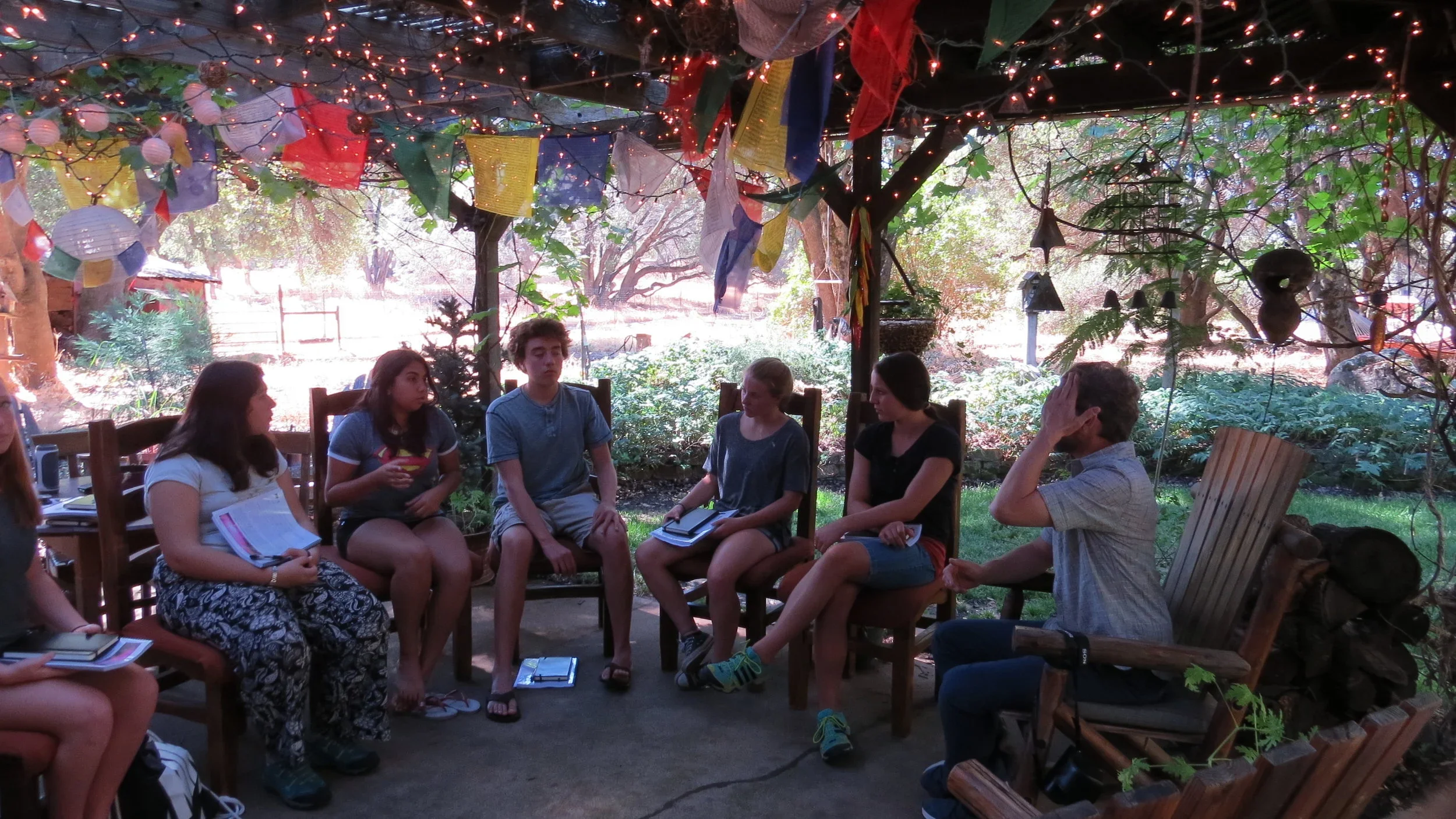 Group of young people sitting in a semi-circle outdoors in a shaded area decorated with colorful banners and string lights, engaged in conversation.