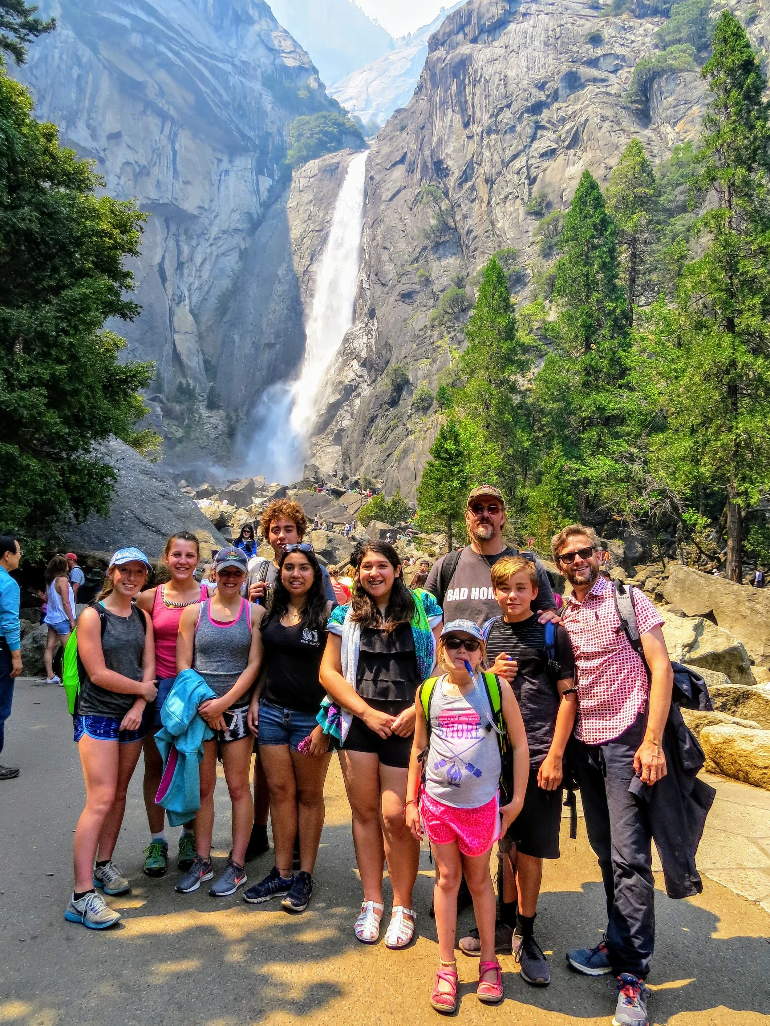 Group of children and adults posing in front of a waterfall in a forested mountain setting.