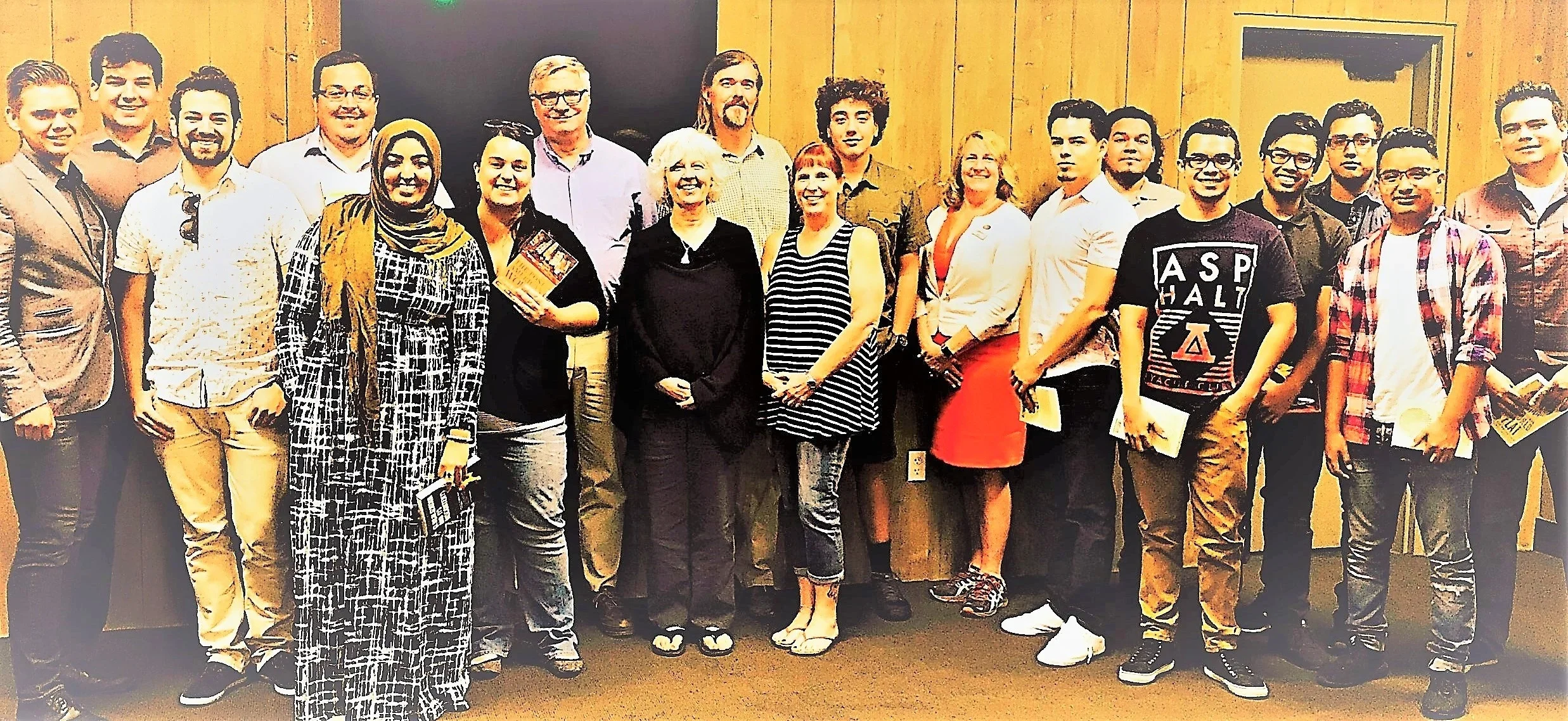 Group of diverse people standing indoors in front of a wooden wall, some holding pamphlets or booklets, smiling for the photo.