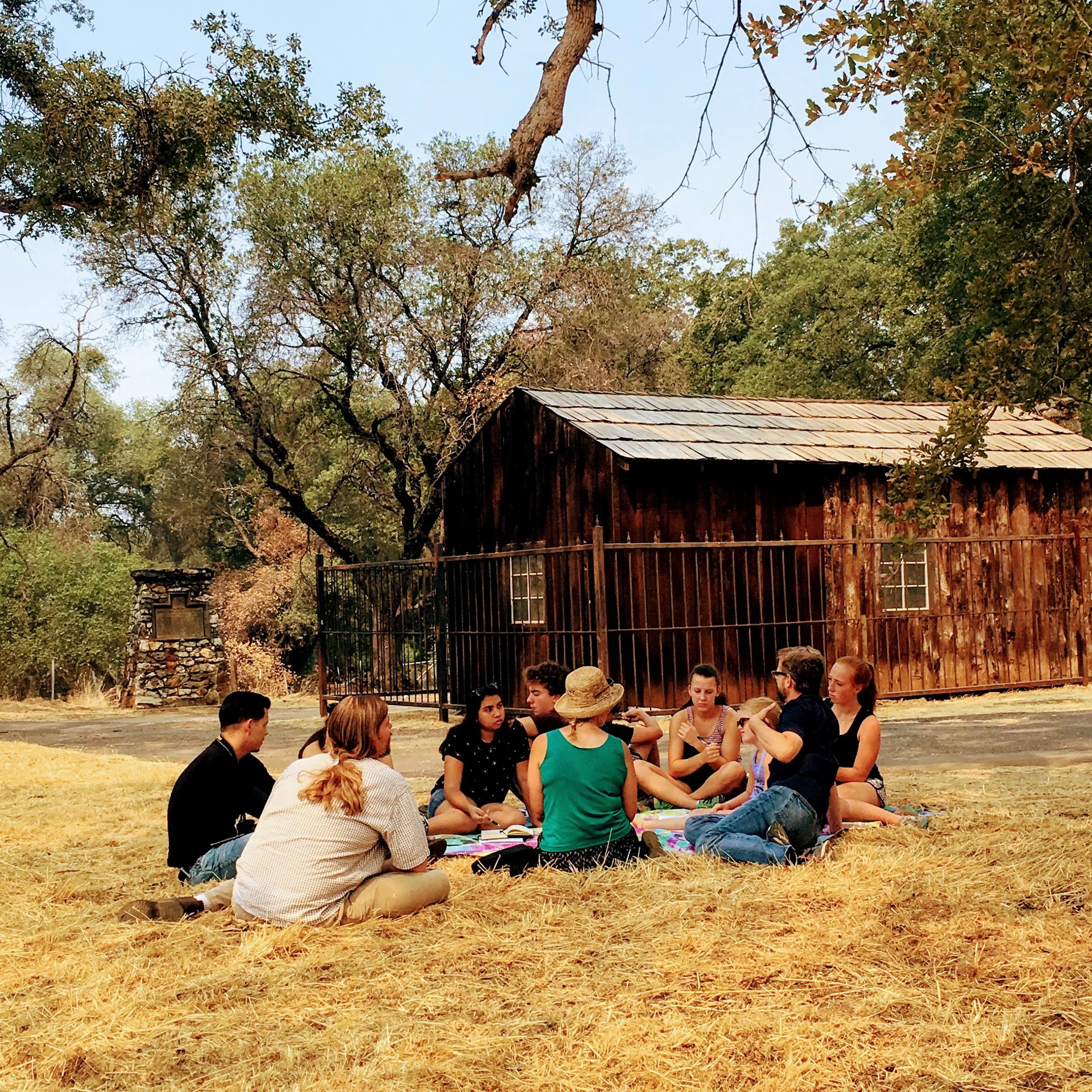 Group of people sitting on a blanket having a discussion outdoors in front of a rustic wooden barn with trees in the background.