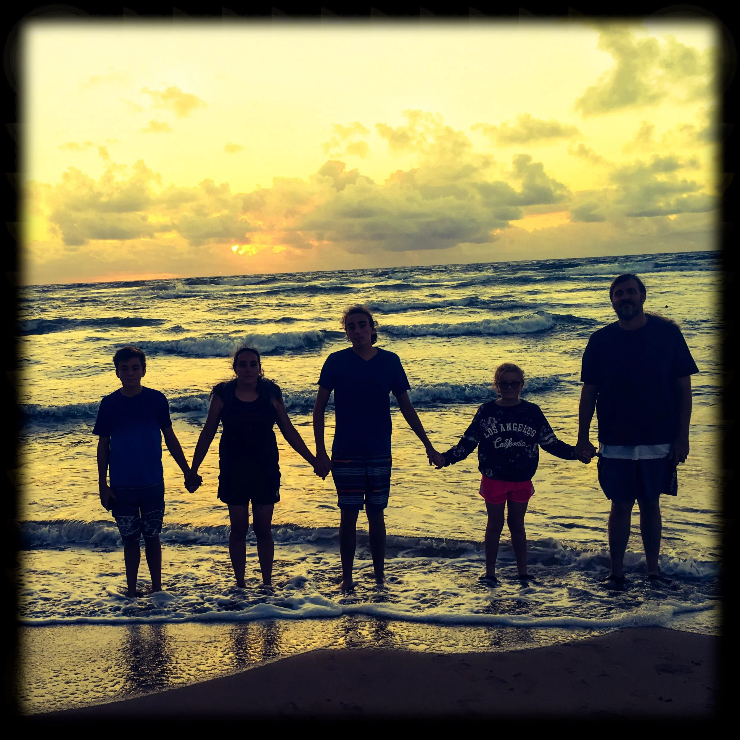 Family holding hands on the beach during sunset with ocean waves in the background.