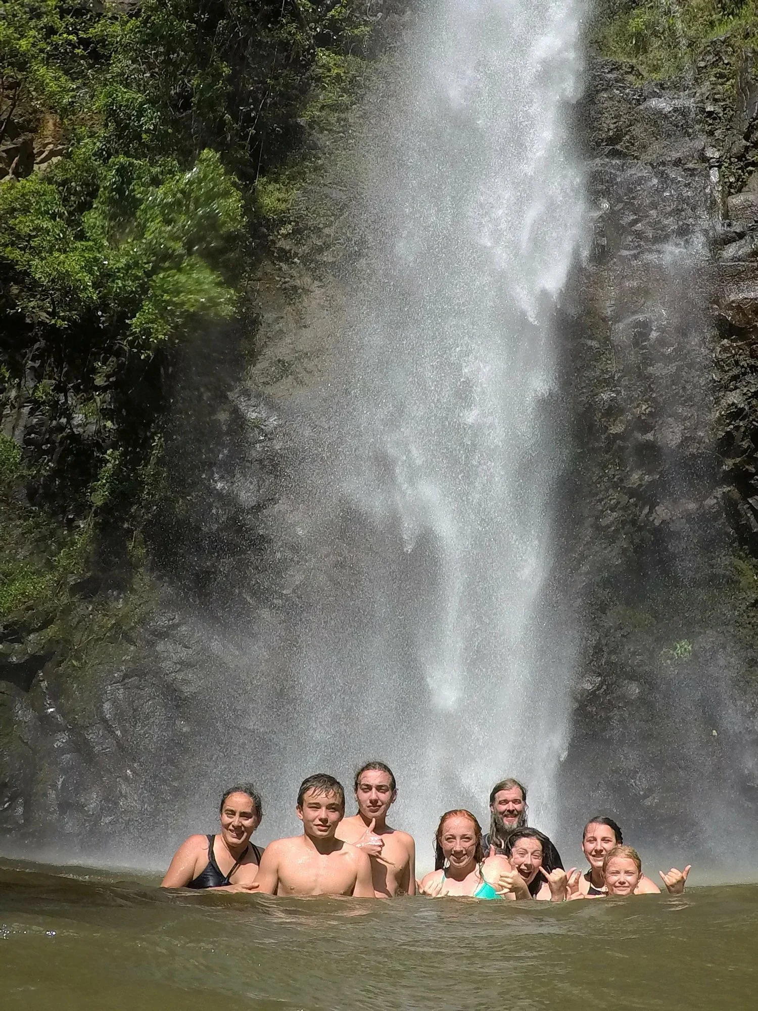 Group of eight people swimming in a waterfall pool, with a tall waterfall and lush greenery in the background.