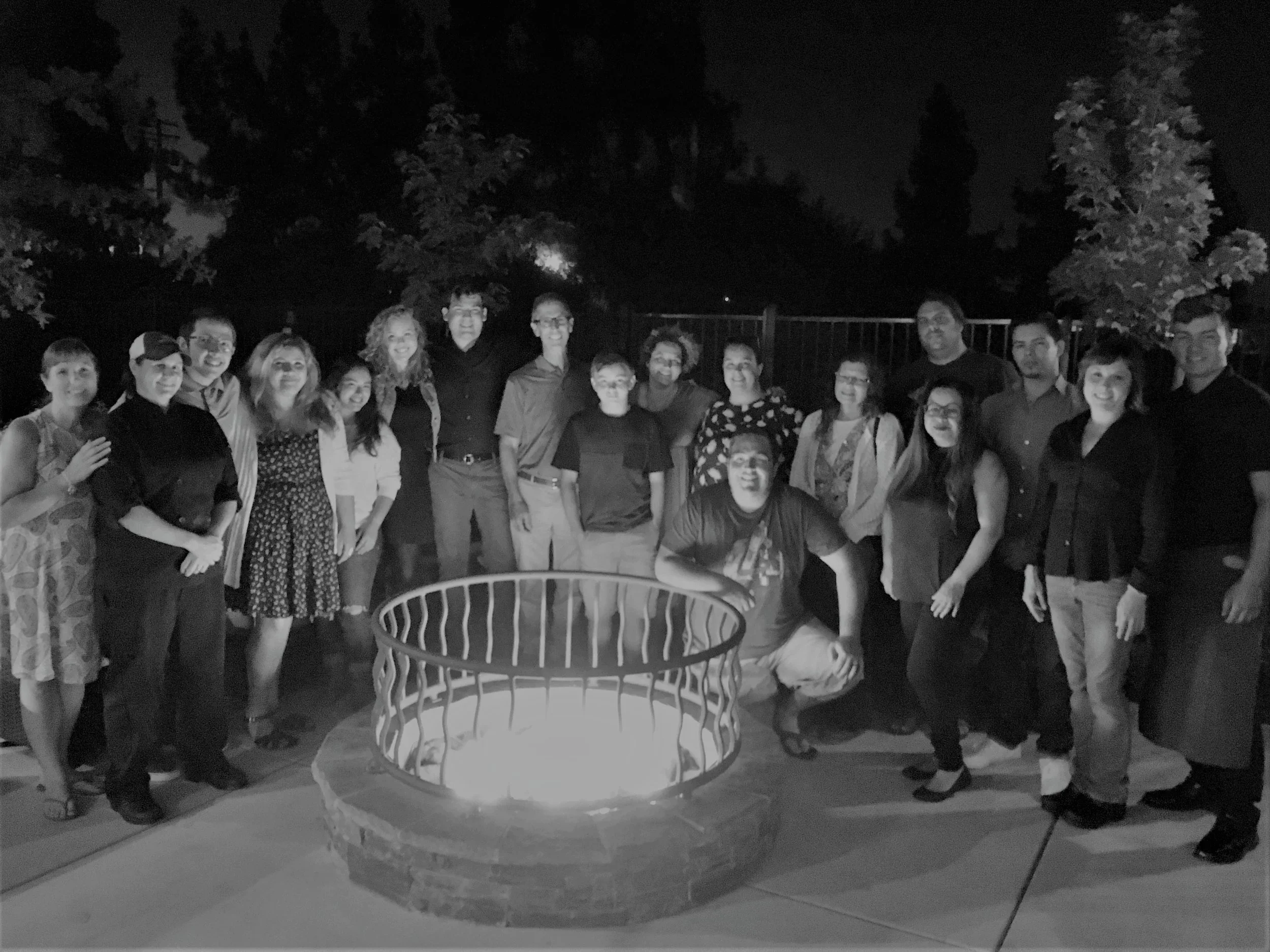 A group of 18 people gathered outdoors at night, standing around a fire pit with a fire inside, smiling at the camera. Trees are visible in the background.
