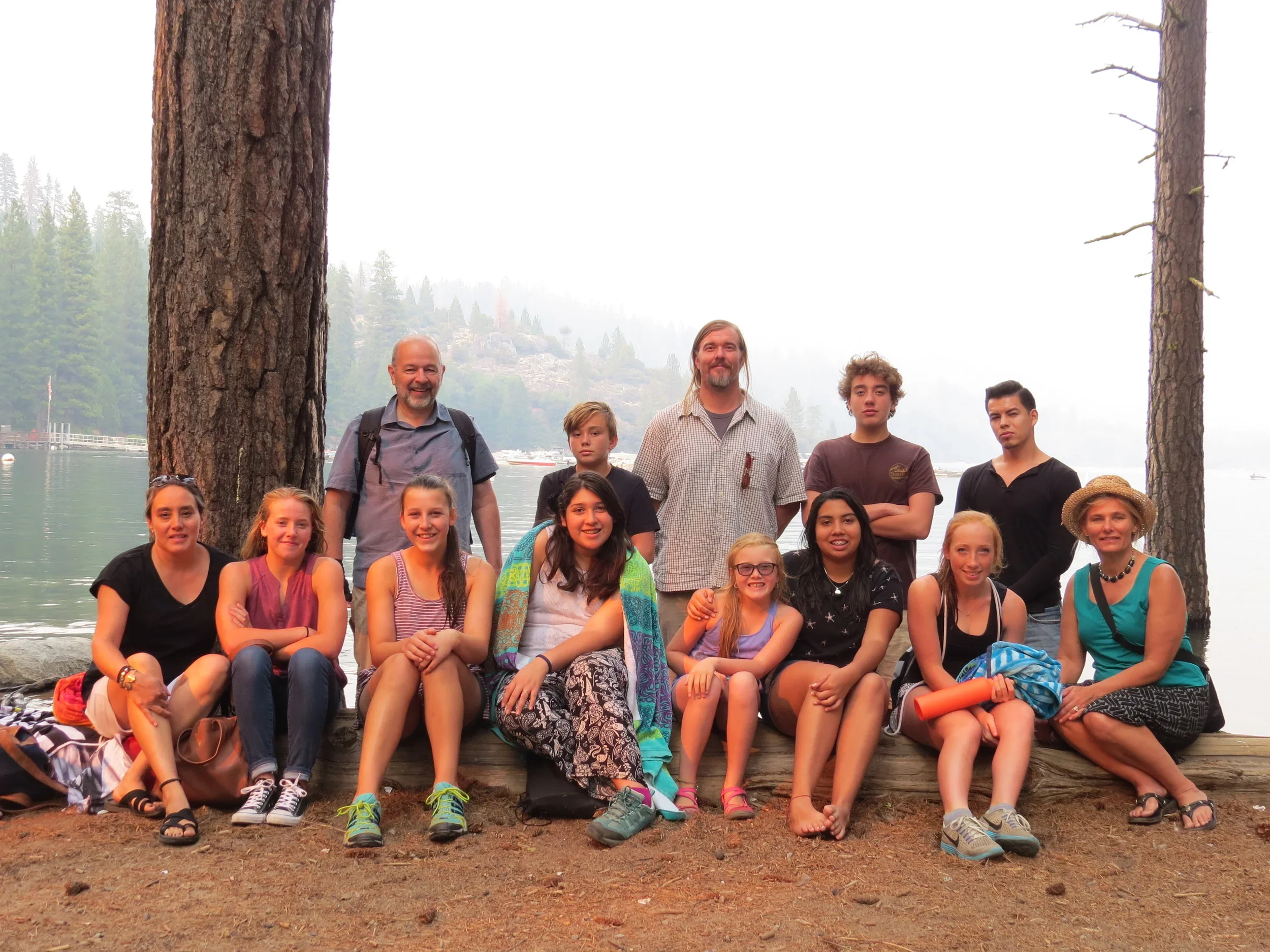 A group of fifteen people, including children and adults, outdoors near a lake with trees and fog in the background. They are sitting and standing on a log and ground, smiling for a photo.