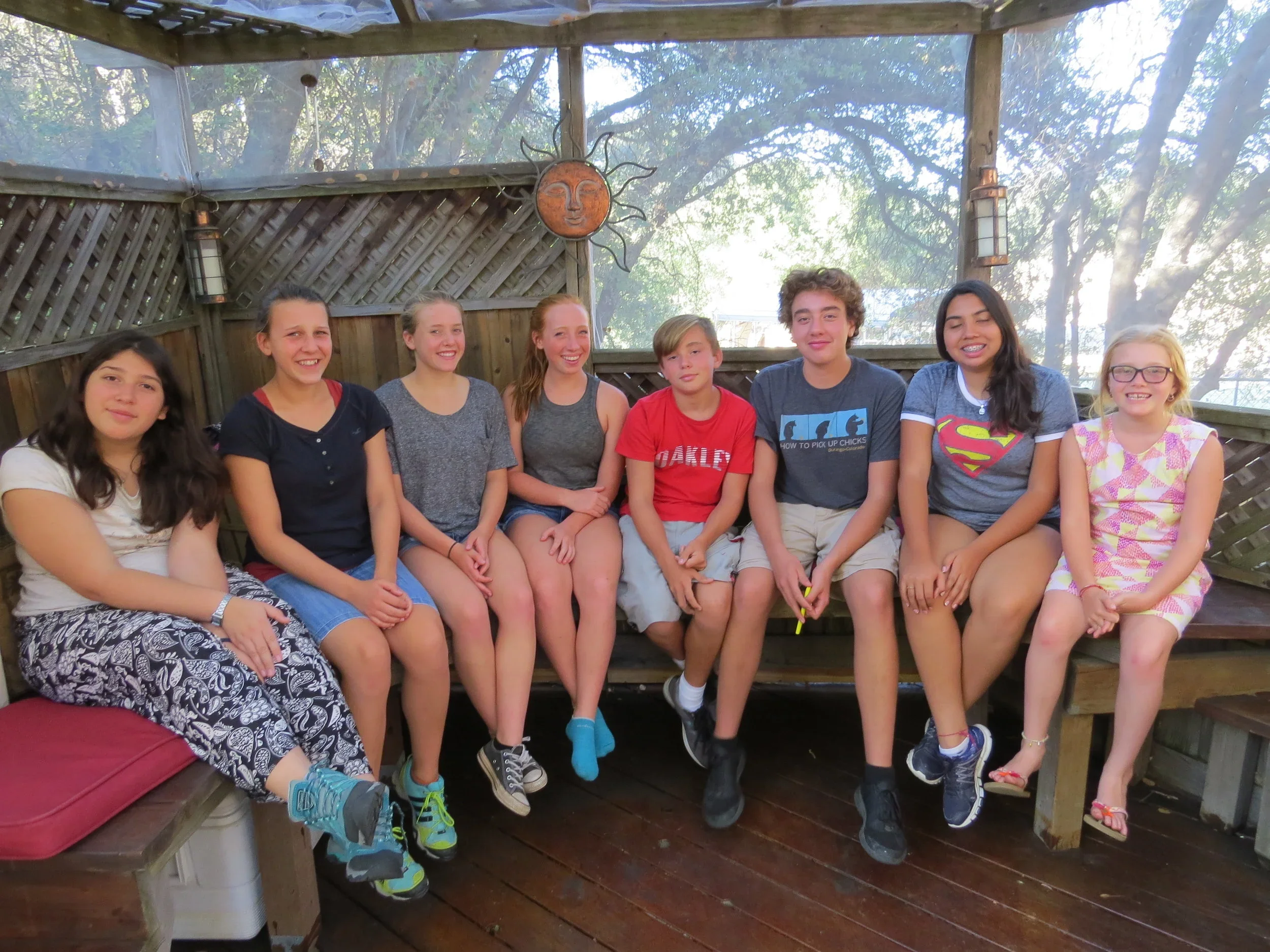 Group of nine teenagers sitting on a wooden bench and patio in an outdoor setting, smiling at camera with trees in the background.