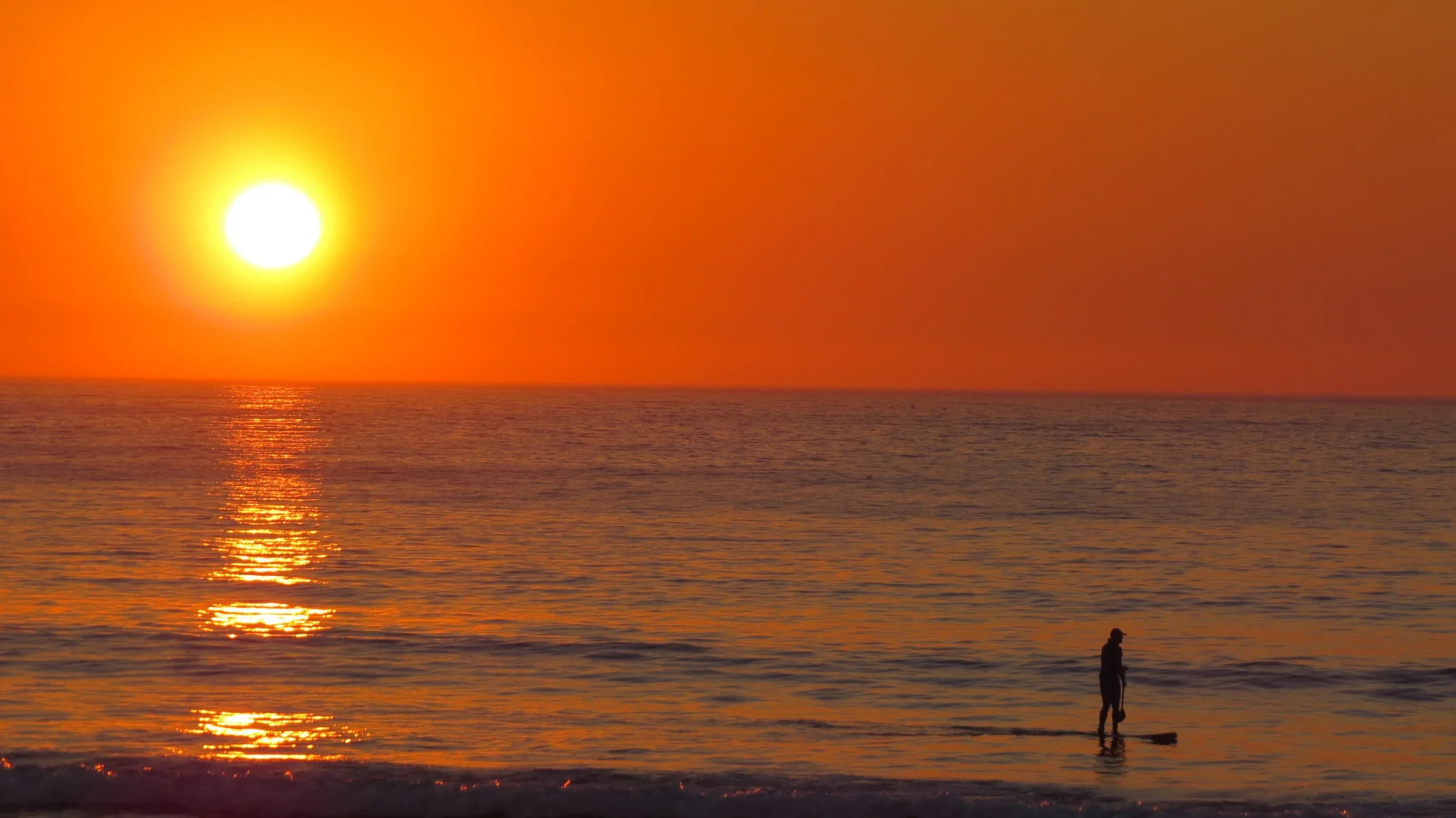 A person standing on a surfboard in shallow water at the beach during a vibrant sunset, with the sun low on the horizon and colorful sky reflected on the water.