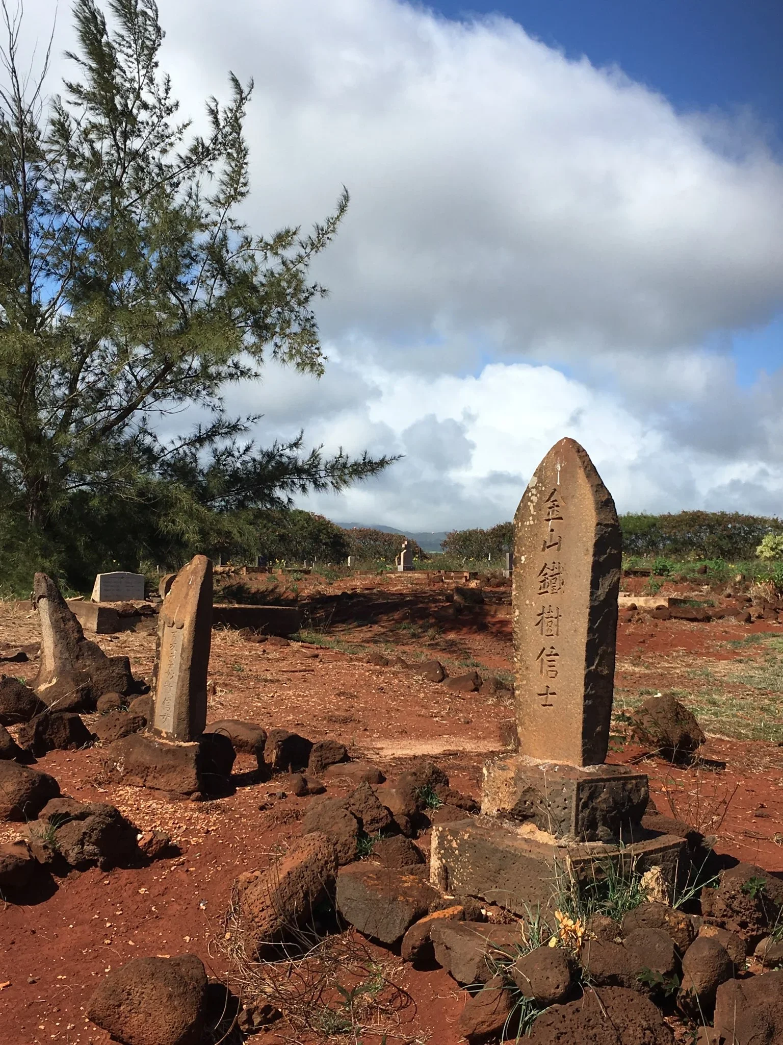 A rocky area with two upright stone monuments with Japanese inscriptions, red dirt ground, scattered rocks and a tree on the left, under a partly cloudy sky.