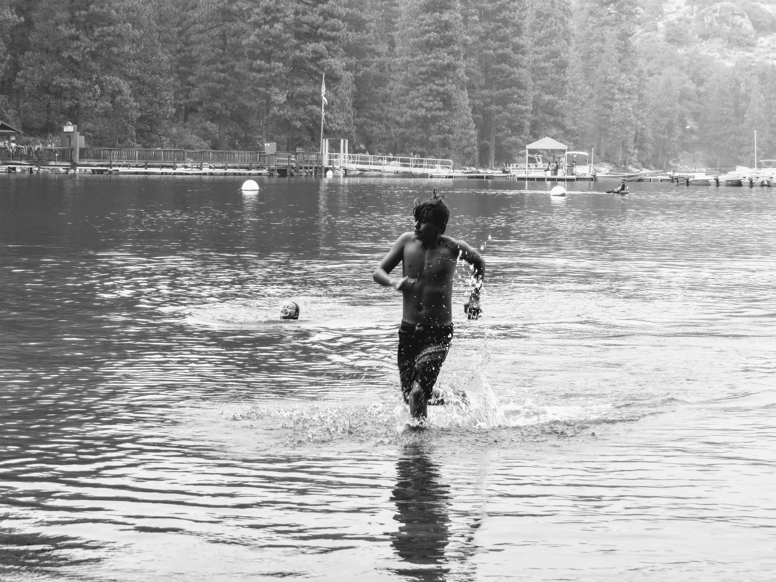 Two children playing in a lake; one is swimming in the background, and the other is running out of the water in the foreground, surrounded by trees and boats at a dock in the distance.