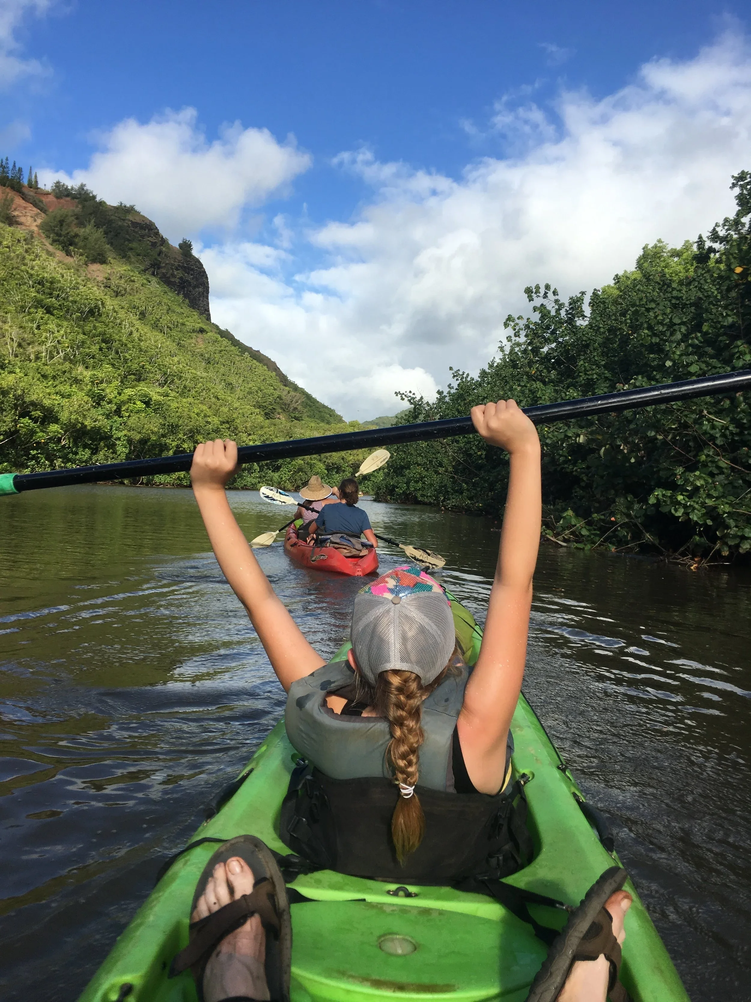 People kayaking on a river surrounded by lush green trees and hills under a partly cloudy sky, with a girl in the foreground holding a paddle above her head.