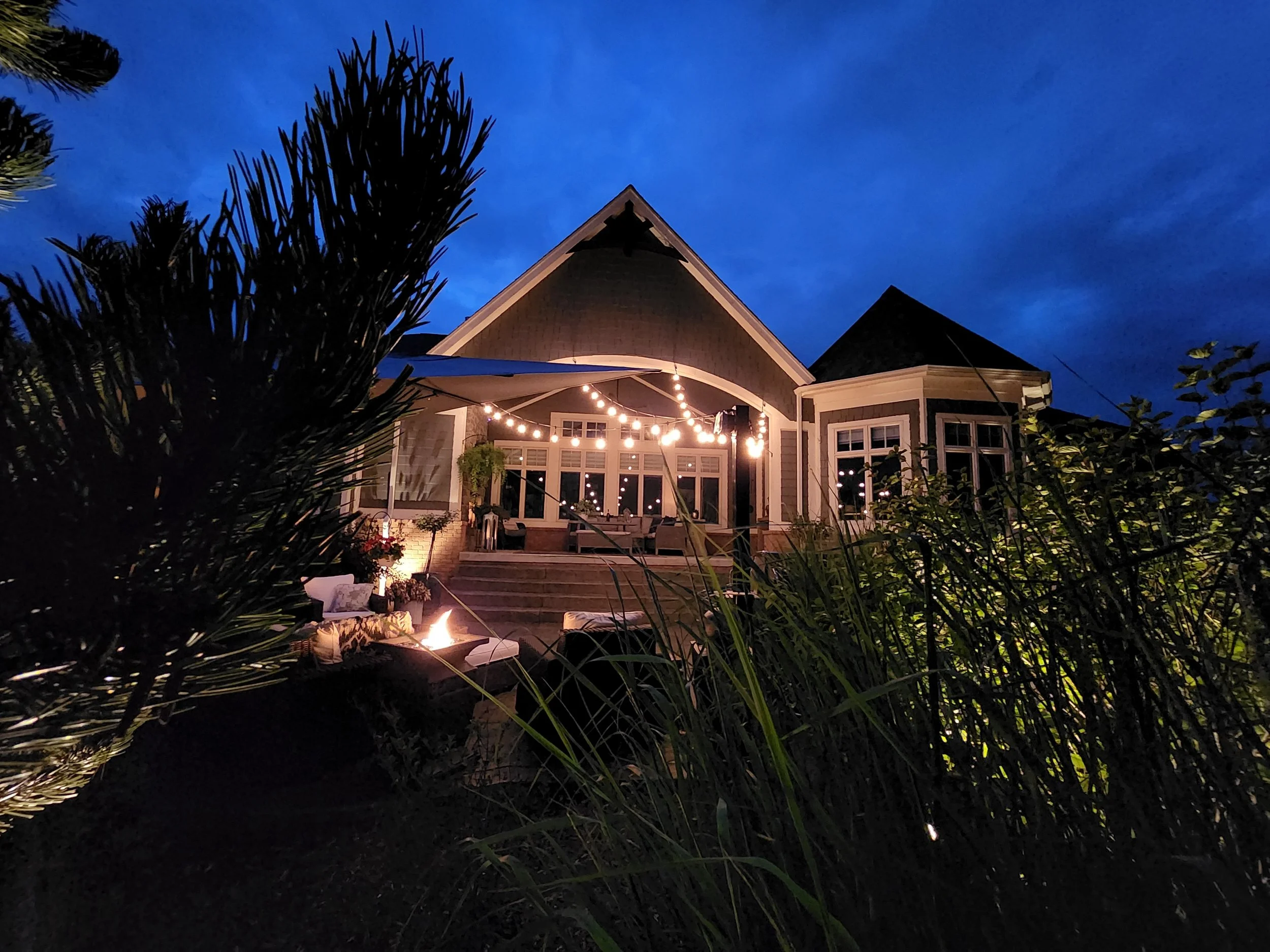 A house with a large porch illuminated by string lights during dusk, surrounded by bushes and trees.