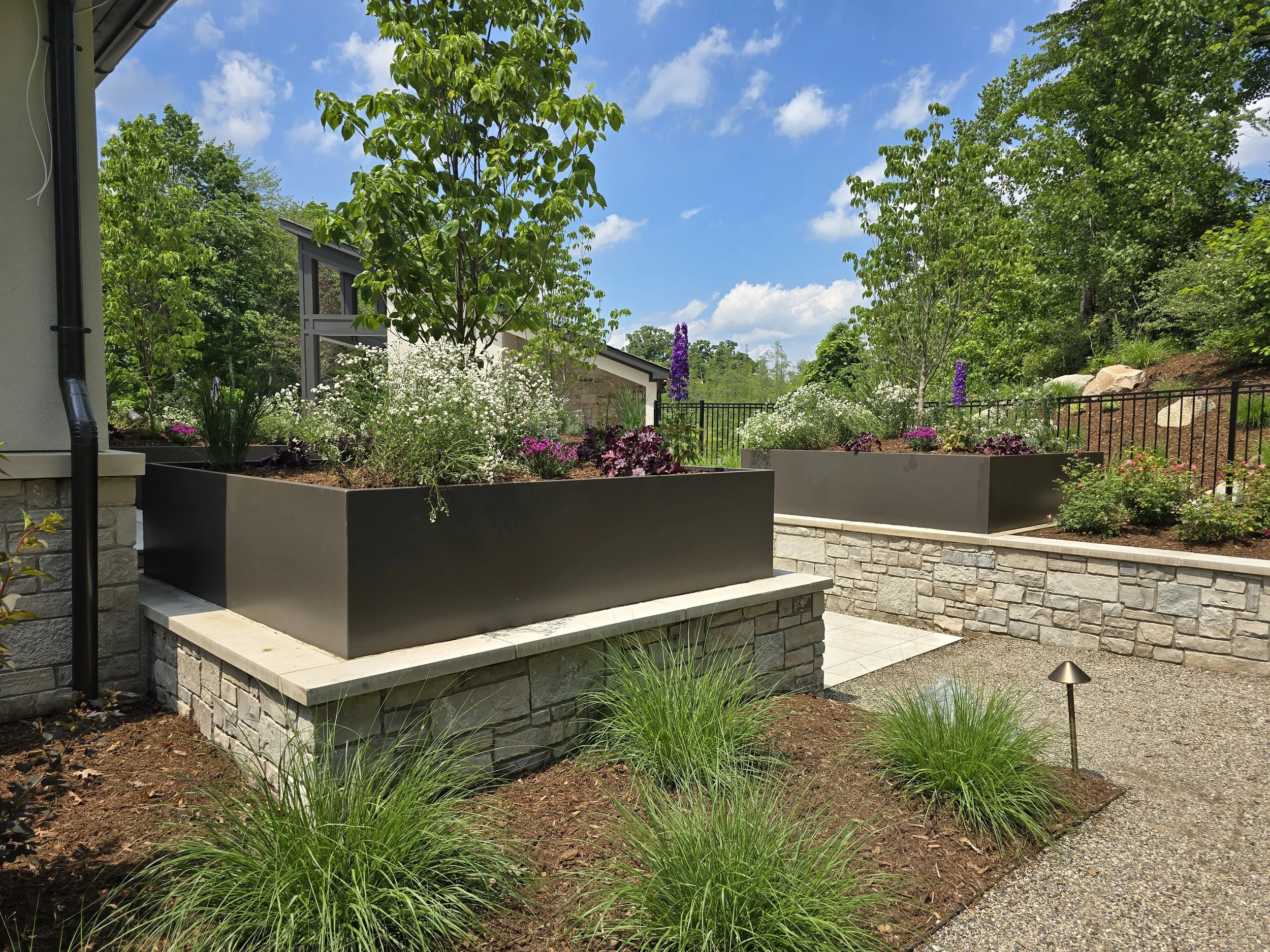A landscaped backyard garden with raised flower beds, lush green trees, and flowering plants under a blue sky with a few clouds.